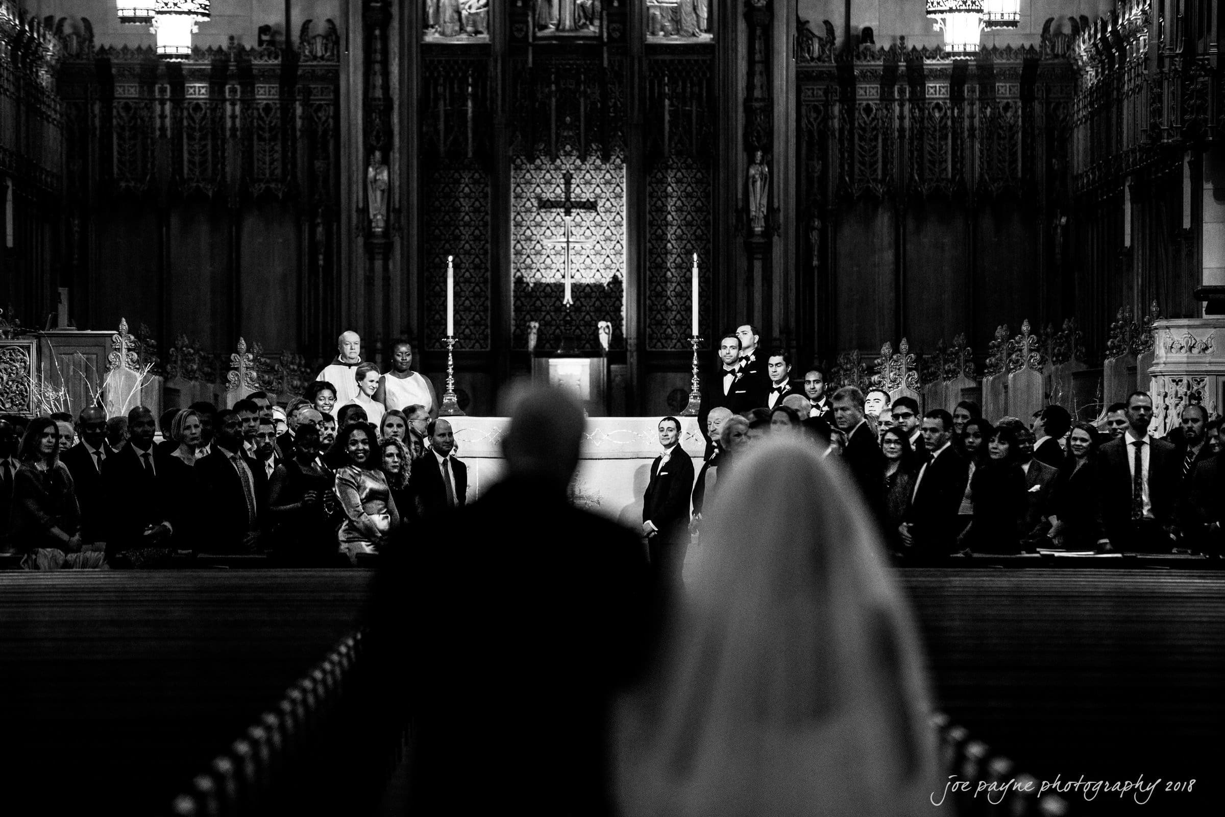 A black-and-white image of a grand church interior, capturing a bride and groom walking down the aisle towards a gathered group of guests and a priest at the altar, with ornate architectural details in the background.