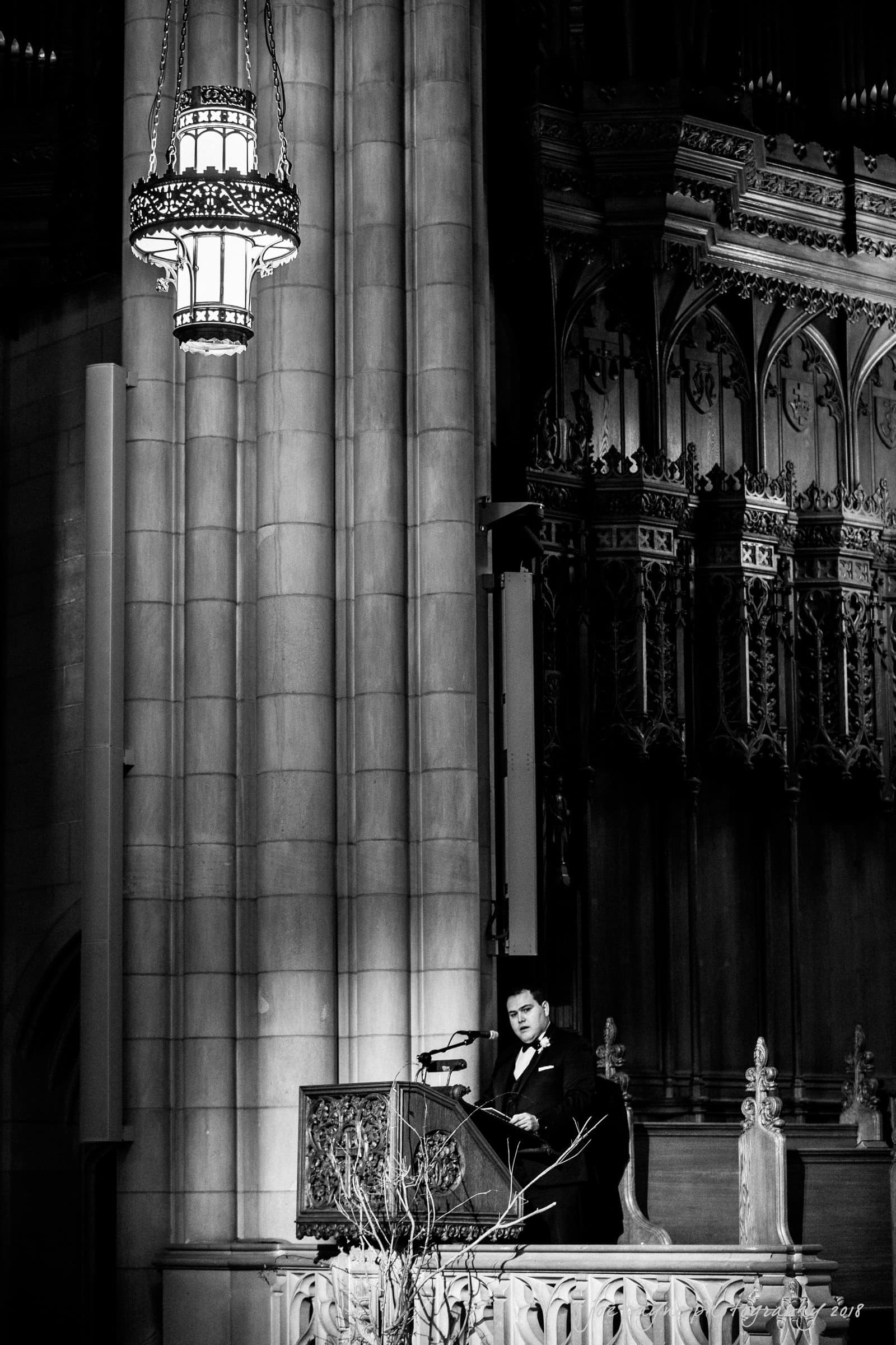 A man stands at a wooden pulpit in a cathedral-style setting, illuminated by a large ornate hanging lamp. The background features intricately carved stonework and archways. The scene is in black and white.