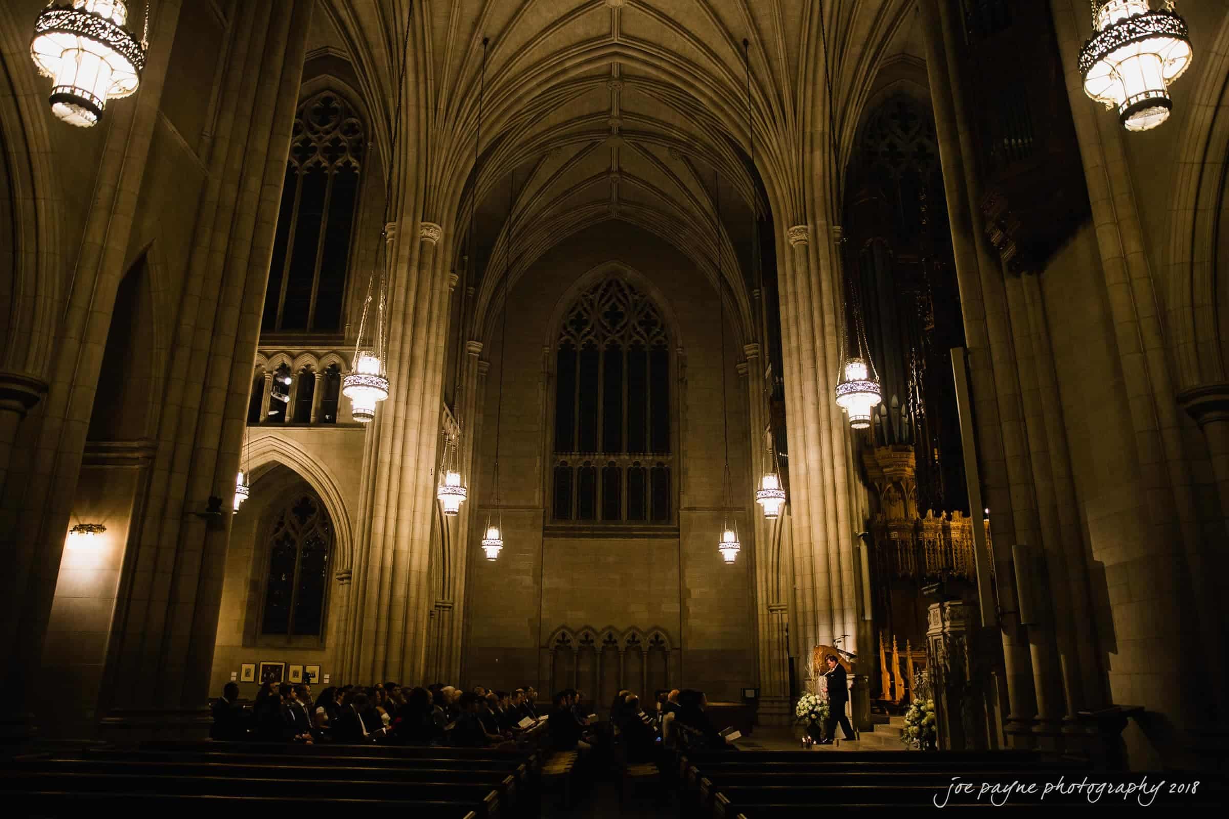 Interior of a grand cathedral with high vaulted ceilings and ornate chandeliers. A wedding ceremony is taking place at the altar, surrounded by seated guests. Soft, warm lighting adds a serene atmosphere to the scene.