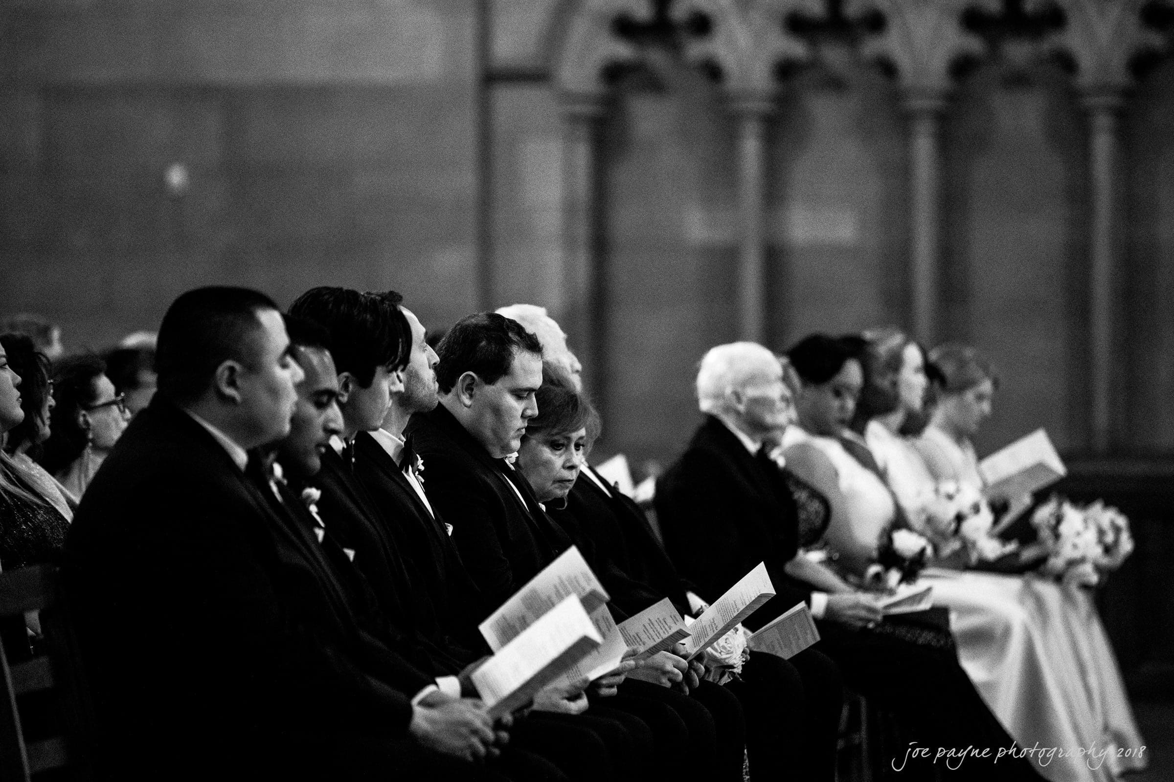 A black and white photo of a wedding ceremony. People are seated in rows, holding programs. They are dressed formally, with men in suits and women in dresses. The setting appears to be a church, with detailed architectural elements in the background.