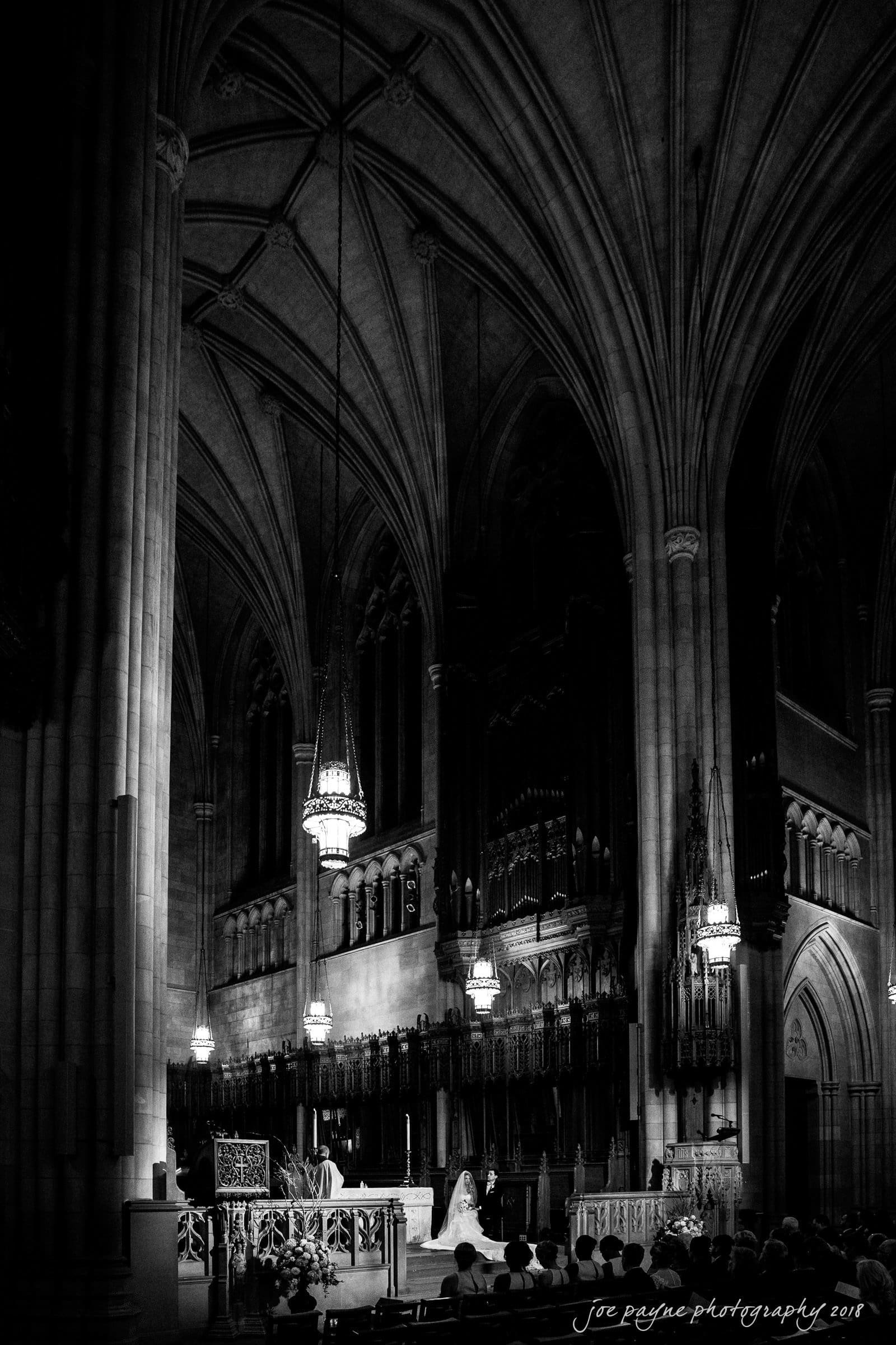 A black-and-white photograph of a grand cathedral interior, featuring high vaulted ceilings and ornate chandeliers. A bride and groom stand near the altar, surrounded by intricate architectural details and gothic elements.