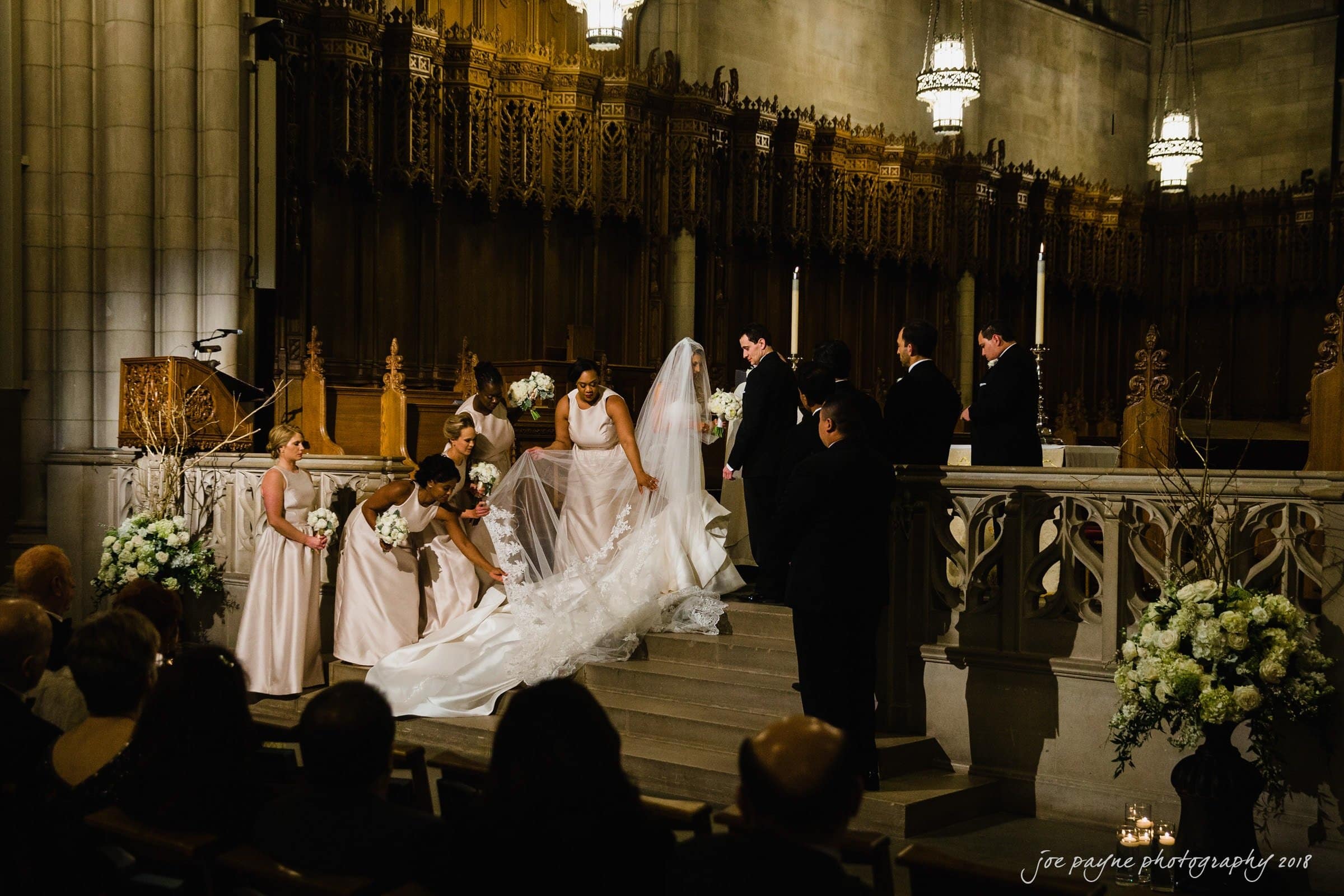 A bride adjusts her gown with the help of bridesmaids at the altar of a church. The groom and groomsmen stand nearby. The setting is ornate with wooden carvings and tall candlesticks, creating a formal and elegant atmosphere.