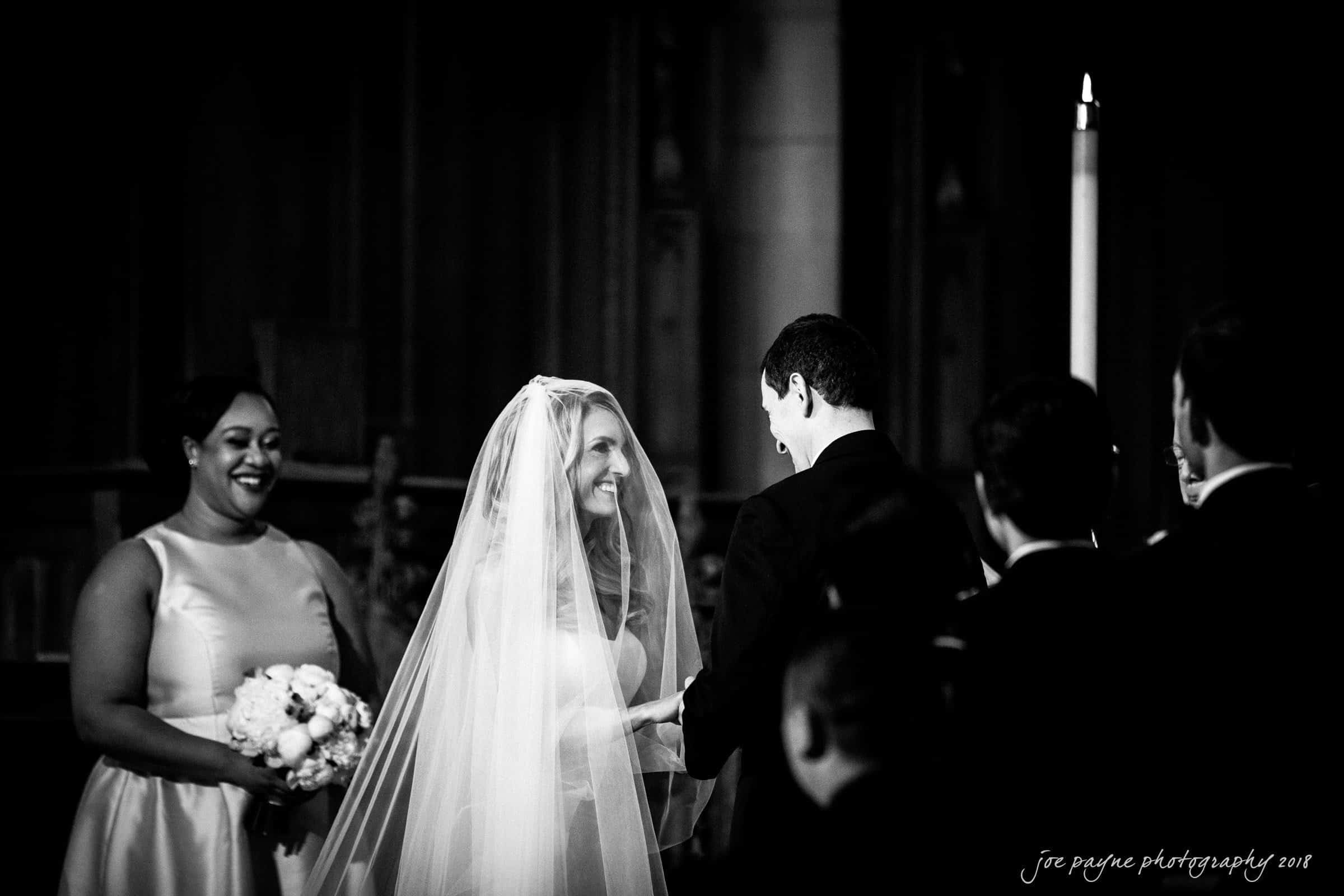 A bride and groom stand during a wedding ceremony, exchanging smiles and vows. The bride wears a veil and the groom is in a suit. A bridesmaid holds a bouquet and watches the couple with a smile. The setting is a dimly lit church.