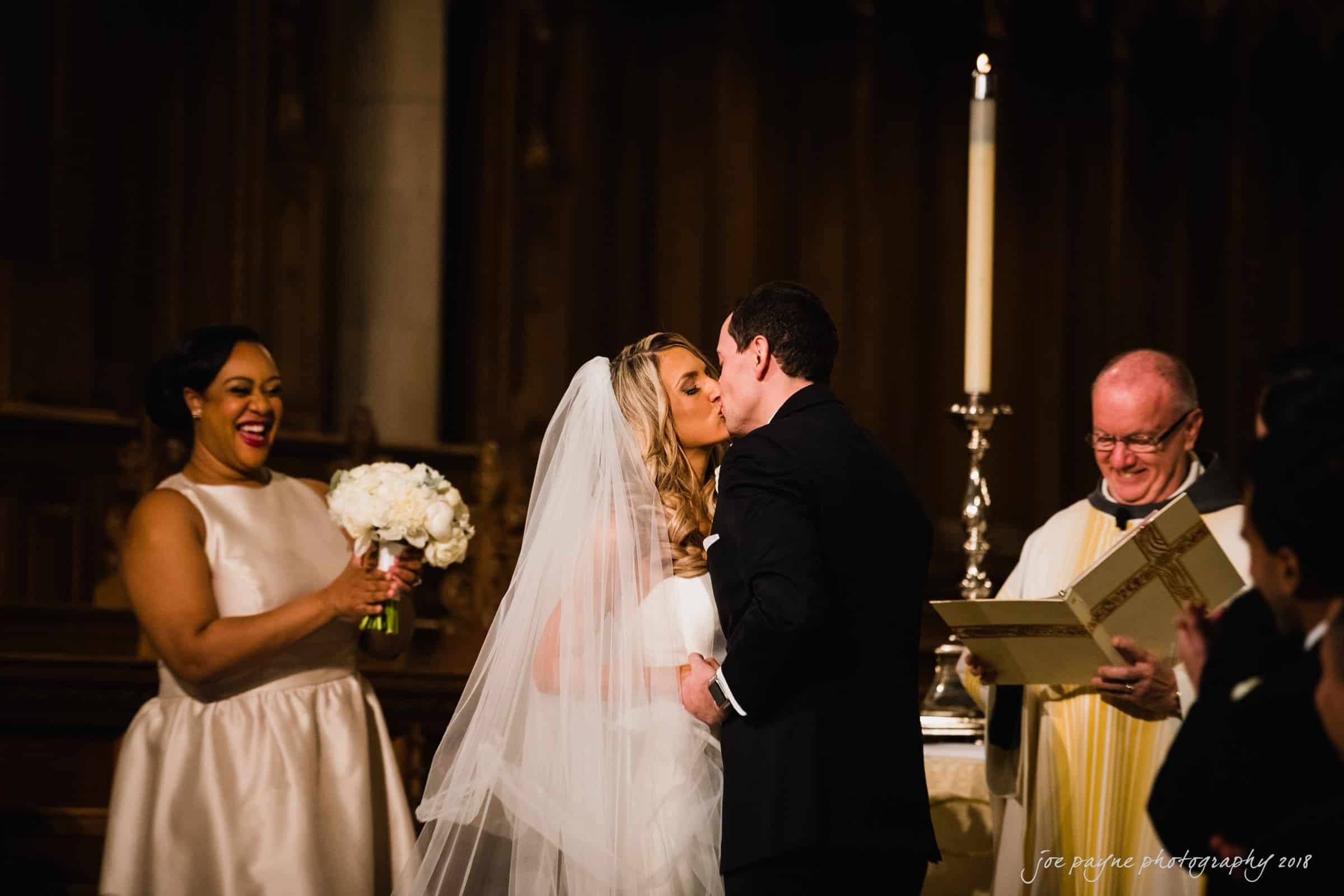 A bride and groom share a kiss during their wedding ceremony. The bride holds a bouquet, while a bridesmaid smiles nearby. A priest stands holding a book, and a large candle is lit in the background.