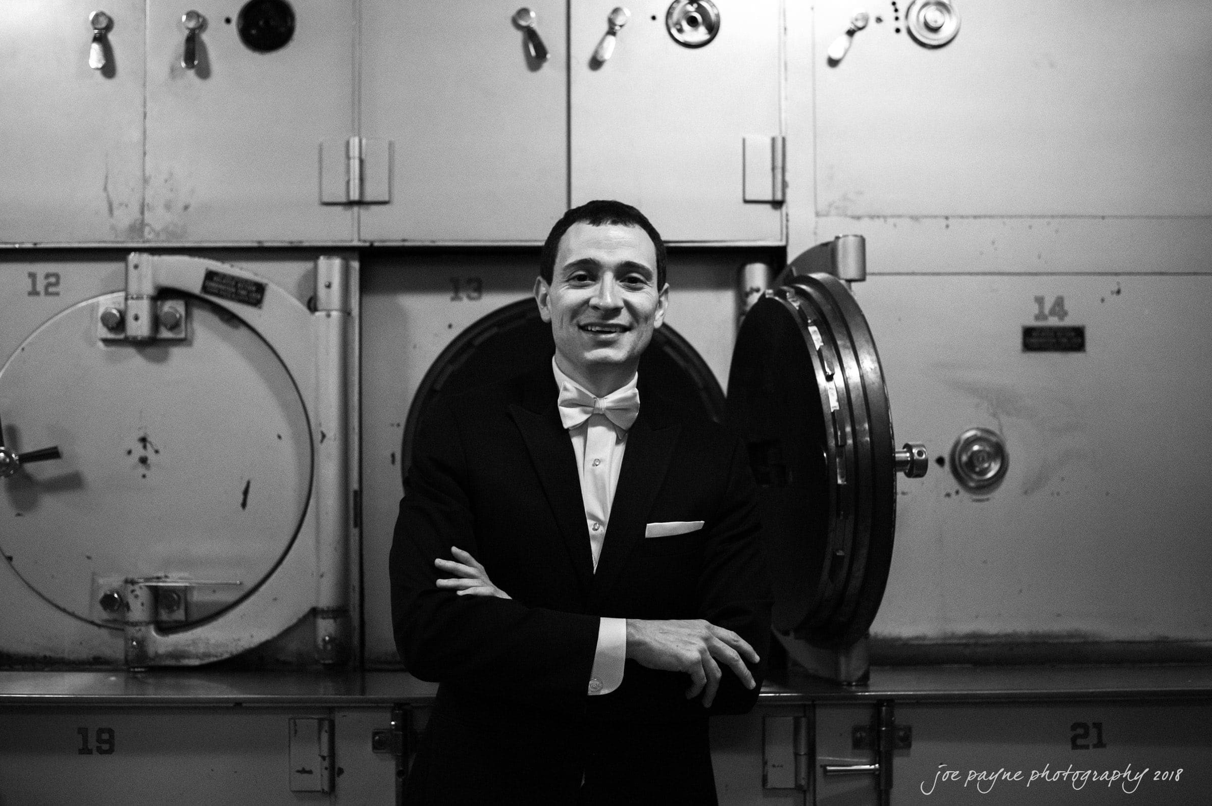 A man in a tuxedo stands smiling with arms crossed. Behind him are industrial machines with round doors, numbered 12 and 21. The setting is black and white, with a vintage, industrial feel.