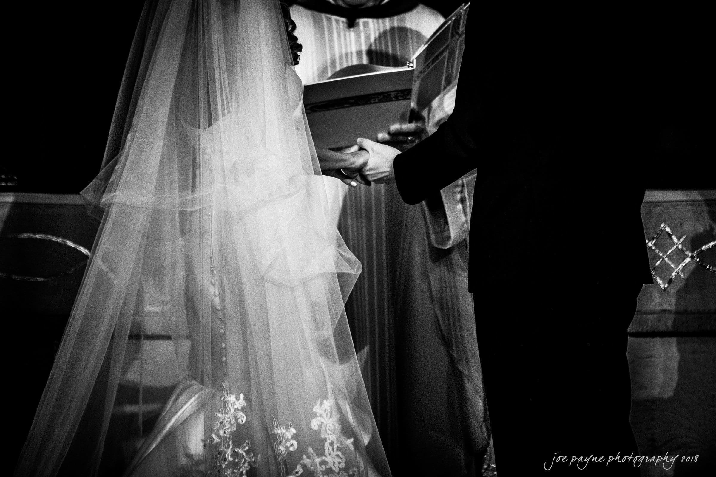 A bride and groom stand together during a wedding ceremony. The bride wears a veil and holds a ring. They face each other, with the groom holding a book. The image is in black and white, capturing an intimate moment.