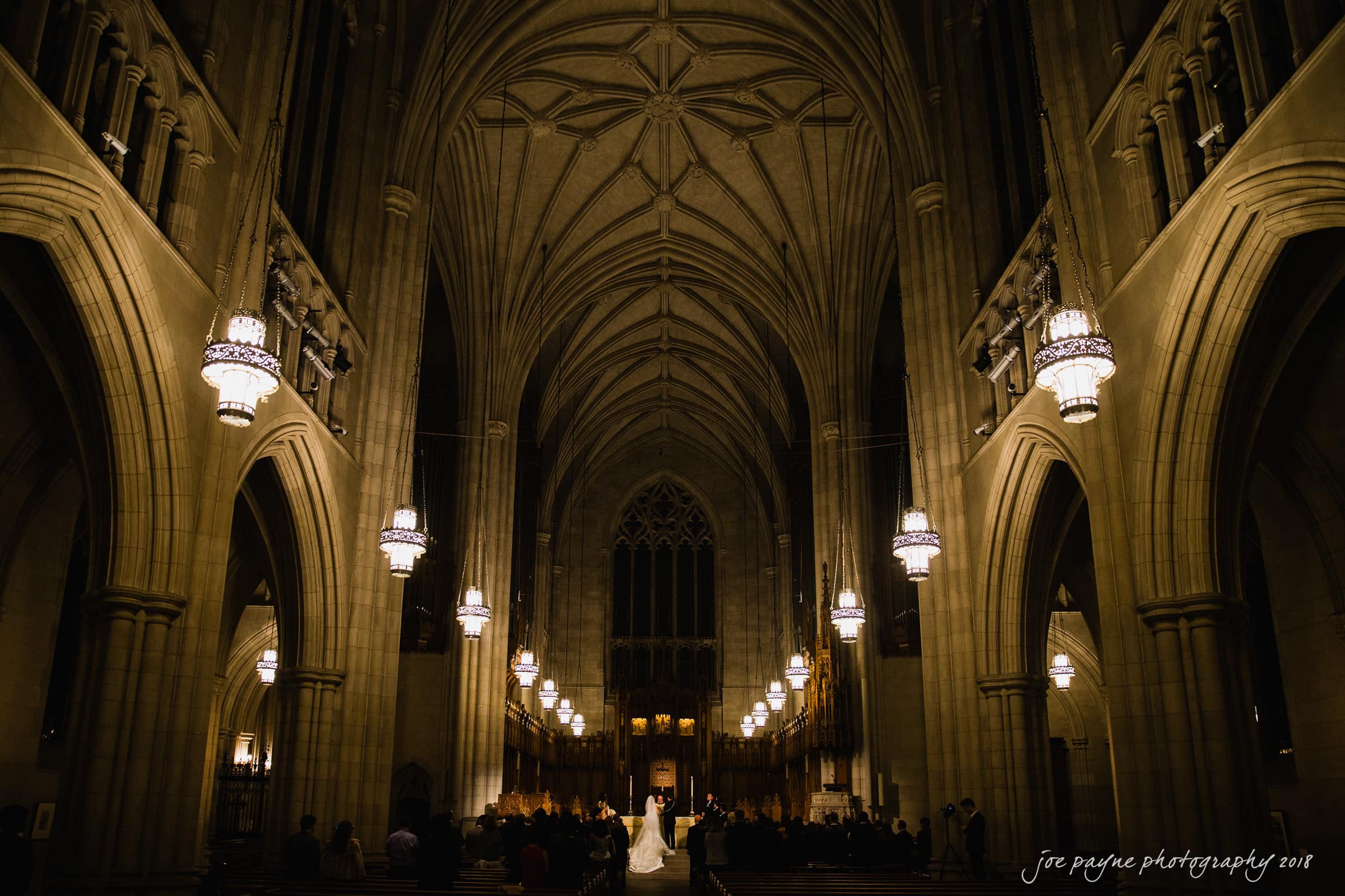 A bride and groom stand at the altar of a grand cathedral, illuminated by soft lighting. Tall arches and intricate architecture surround them, creating a majestic and solemn atmosphere. Guests are seated in the pews, witnessing the ceremony.