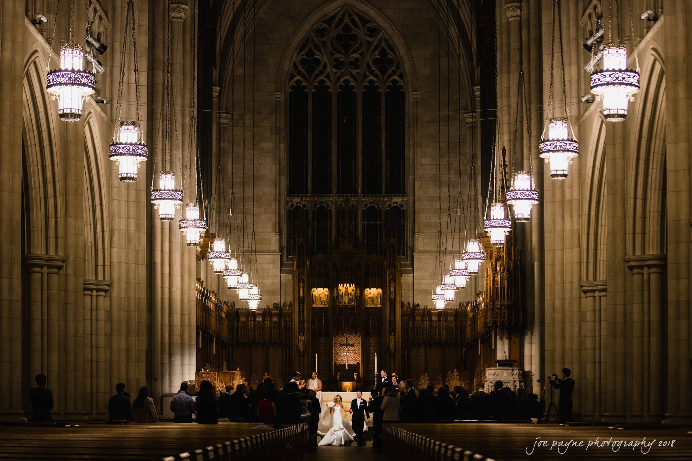 A couple stands at the altar in a grand cathedral during their wedding ceremony. The interior is illuminated by hanging lights, and the arched ceiling and stained-glass windows create a dramatic backdrop. Guests are seated on either side.