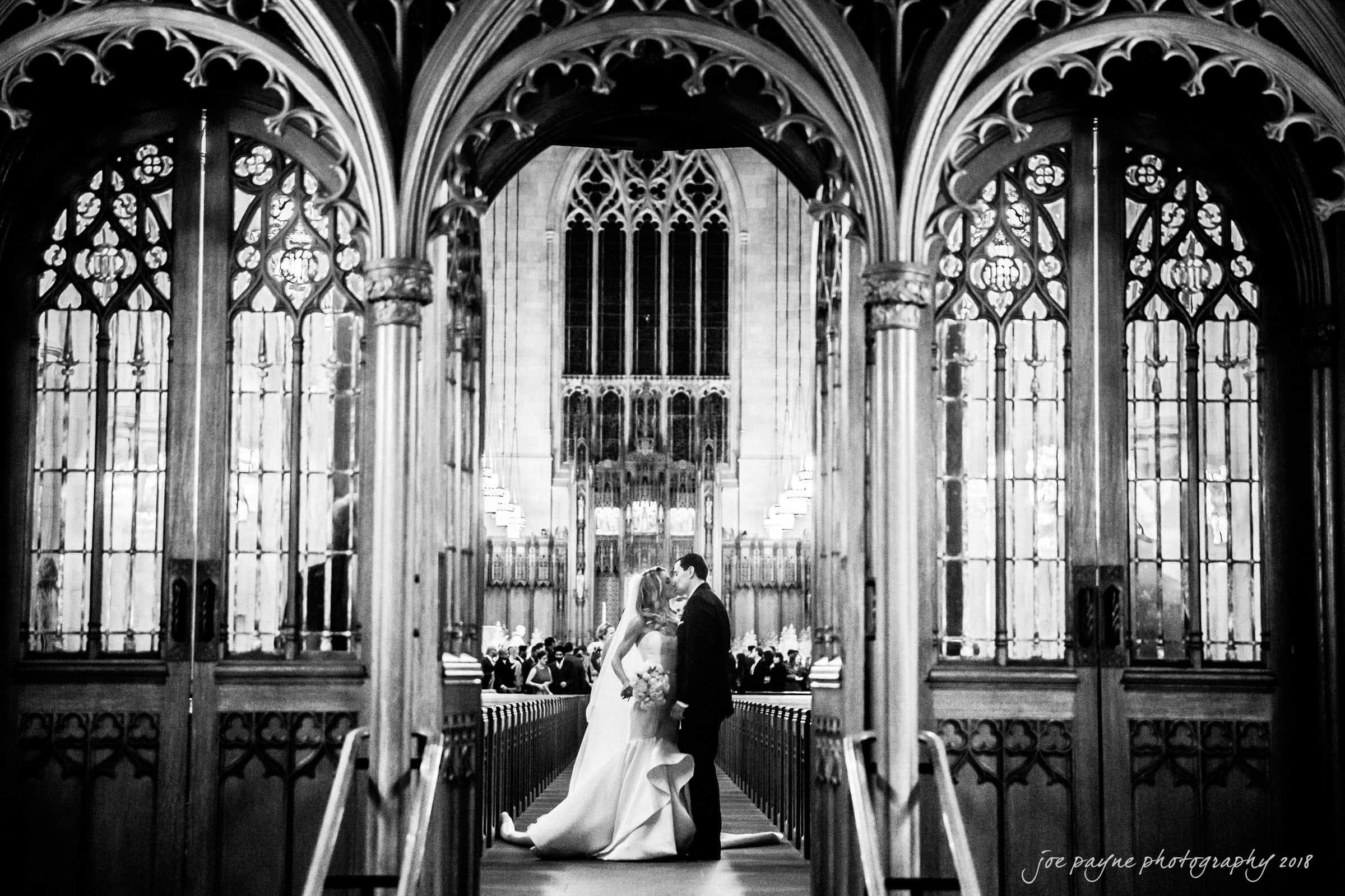 Duke Chapel Weddings A creative wedding photograph at Duke Chapel, shot from the narthex through the ornate wooden doors as the newlyweds, Brandy and Gabe, stop to kiss at the end of their recessional aisle.
