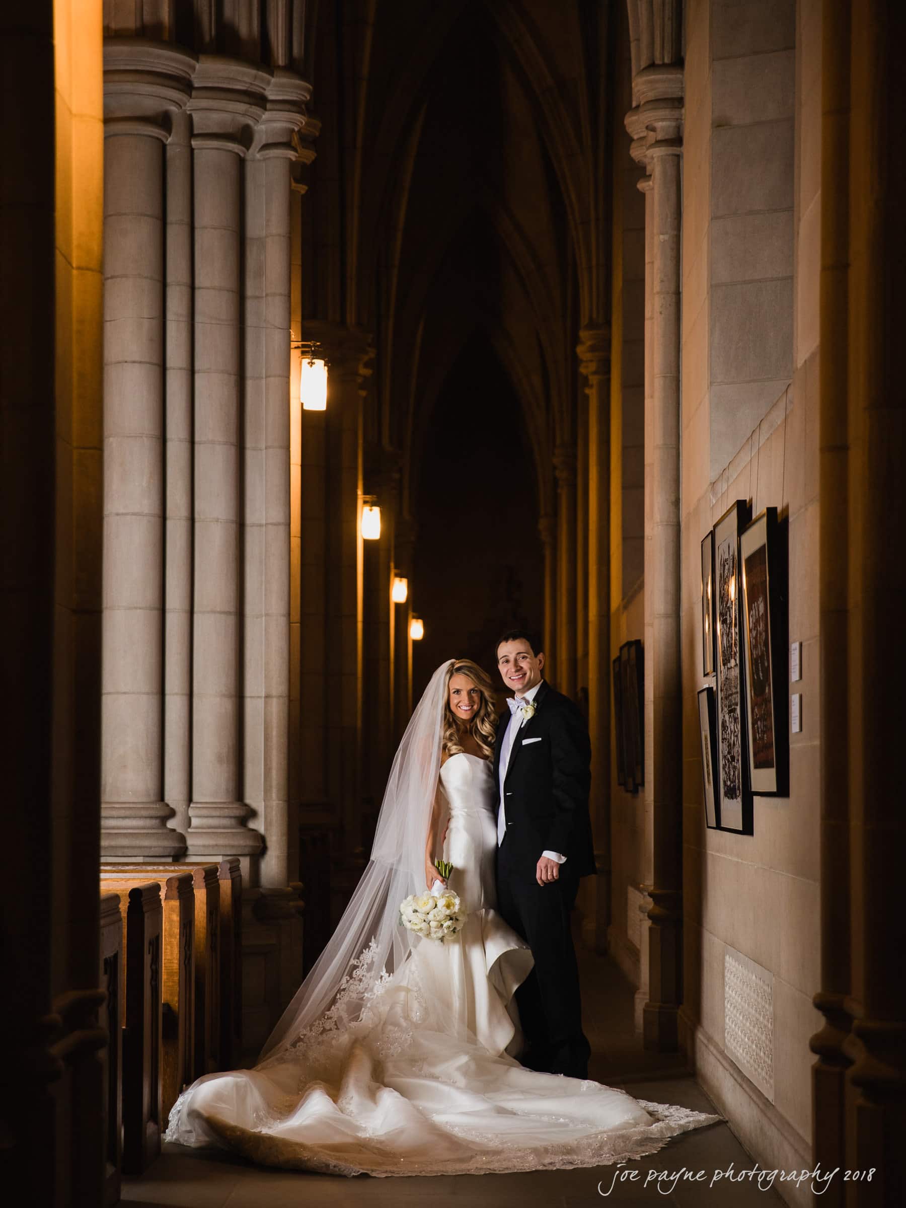 A bride and groom stand in a dimly lit, elegant hallway with tall stone columns and arched ceilings. The bride is in a long white gown with a veil, holding a bouquet, and the groom is in a tuxedo. Warm lights illuminate the scene.