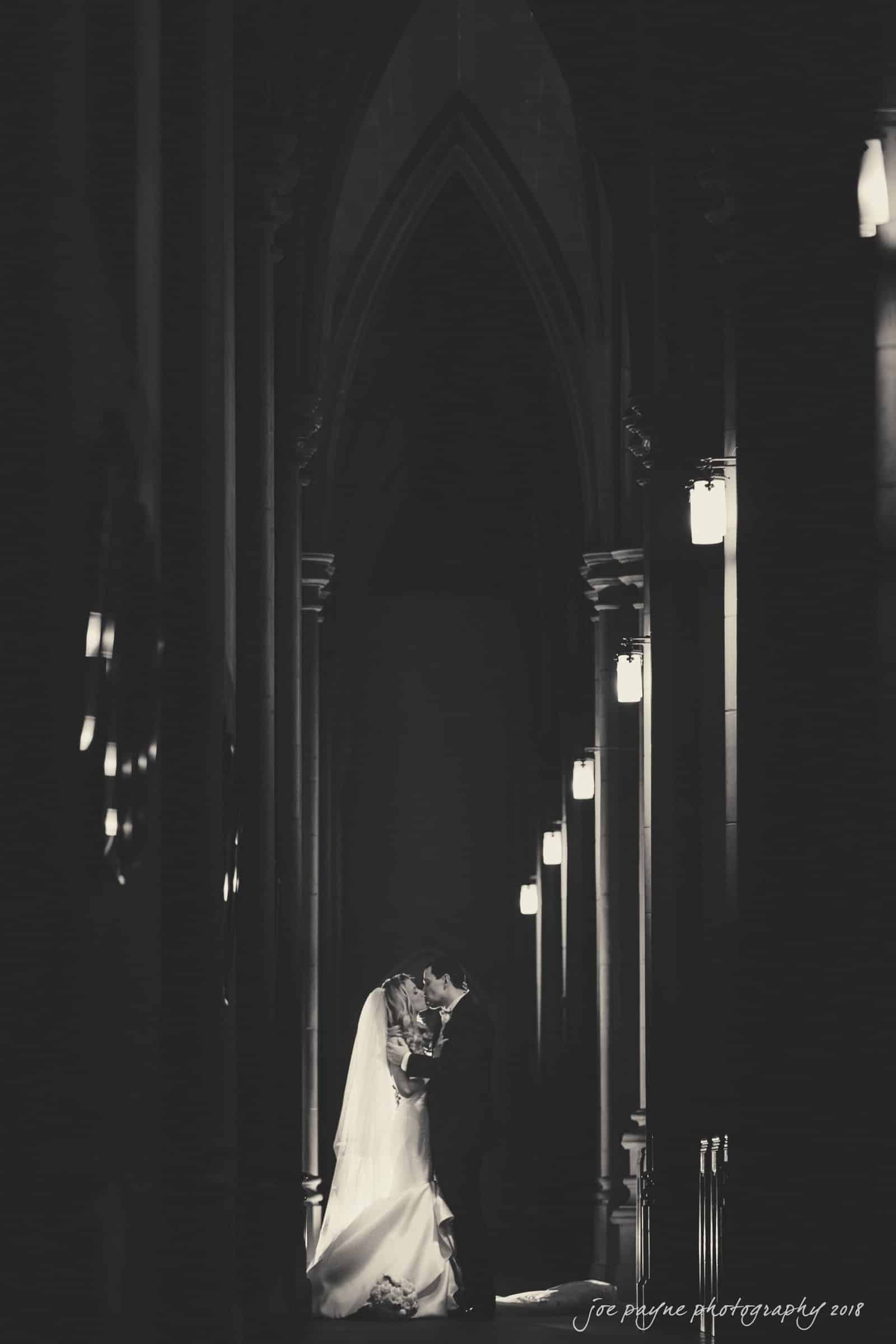 A bride and groom embrace and kiss in a dimly lit cathedral with tall arches. The bride is in a white gown, and the groom wears a suit. Soft lights illuminate the scene, creating a romantic and intimate atmosphere.