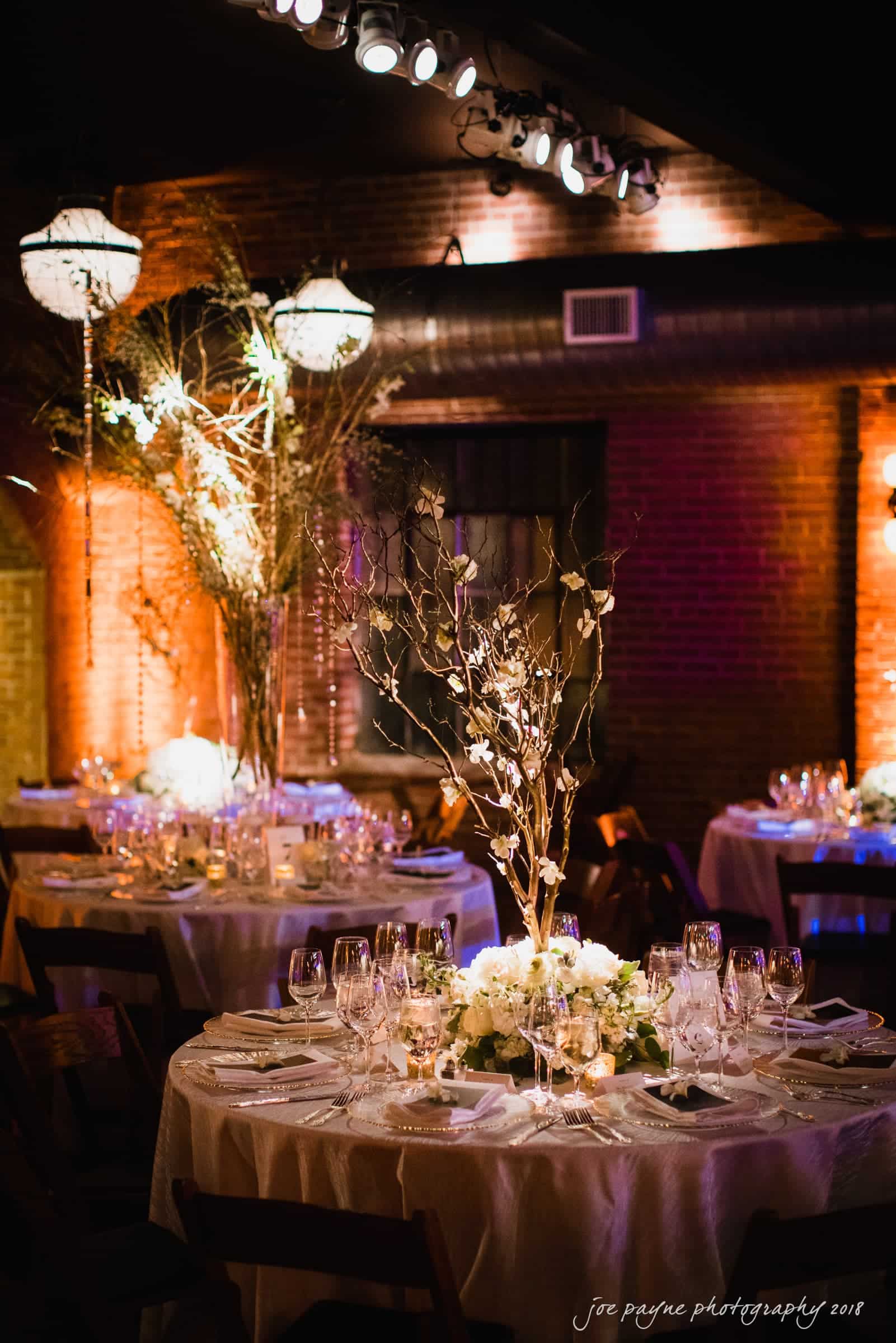 A dimly lit, elegant indoor wedding reception with round tables. Each table is adorned with white floral centerpieces and tall branches. Wine glasses and plates are neatly arranged, and warm lighting creates a cozy atmosphere.