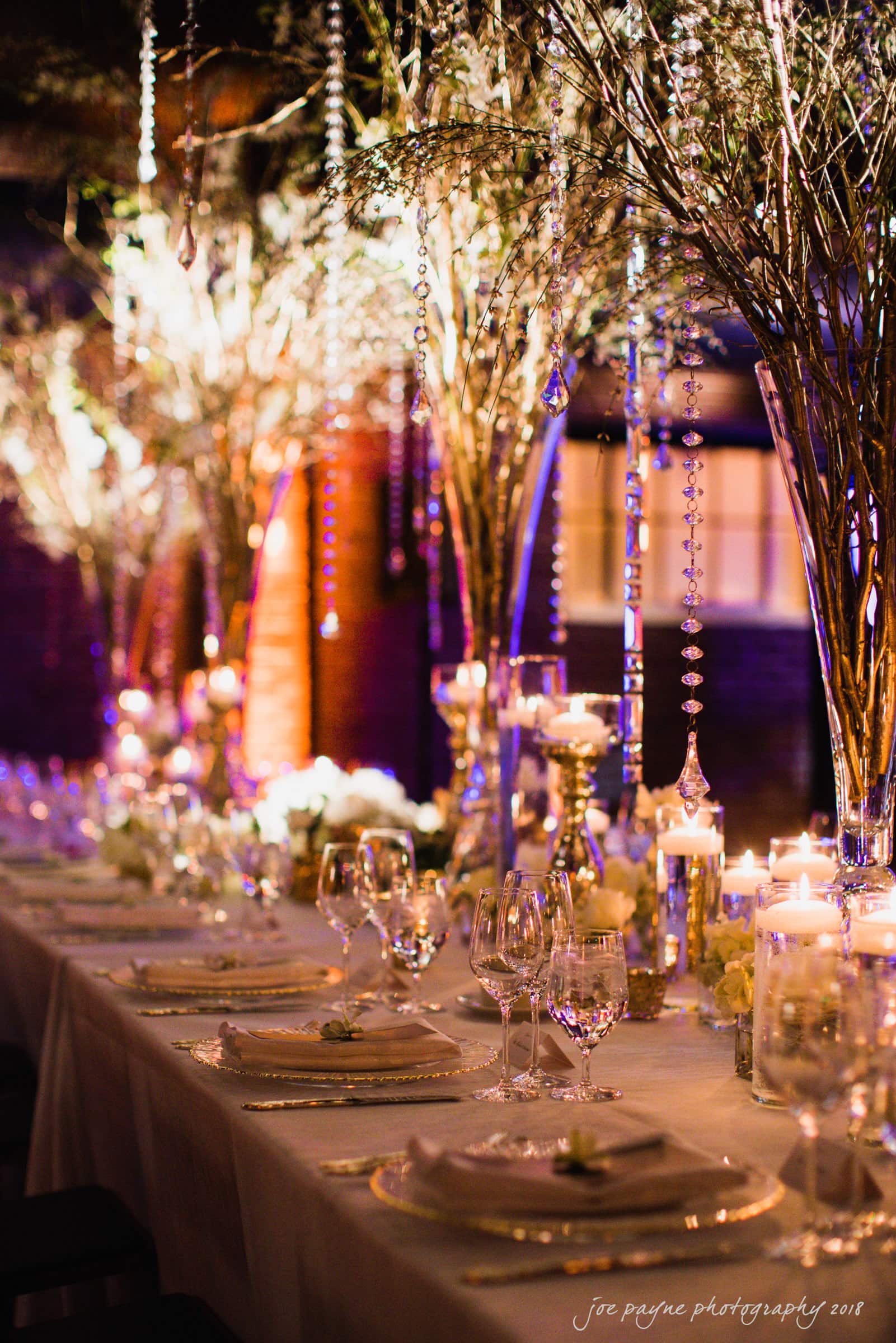 A long, elegantly set dining table with tall, crystal-accented centerpieces and glowing candles, creating a warm, inviting ambiance. The soft lighting and sparkling decorations add a luxurious touch to the setting.