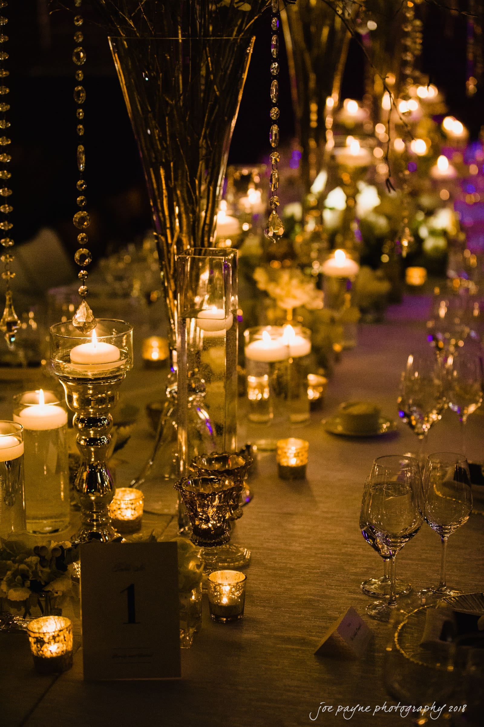 Elegant table setting with tall, ornate candle holders and hanging crystal strands. Lit candles create a warm, inviting glow. The table is adorned with white flowers and sparkling glassware, with a small card displaying the number one.