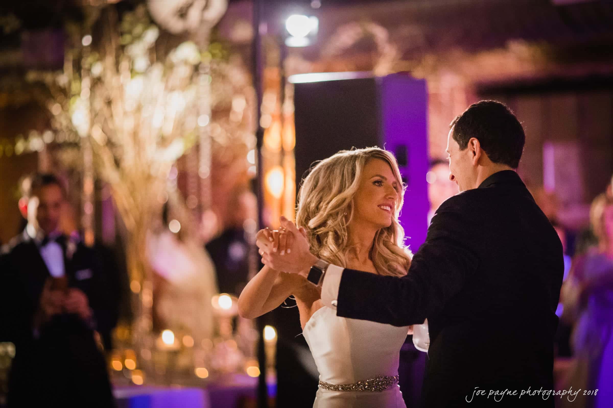 A couple dances under soft lights at a formal event. The woman wears a white dress, and the man is in a suit. The room is warmly lit, with decor in the background creating an elegant ambiance.