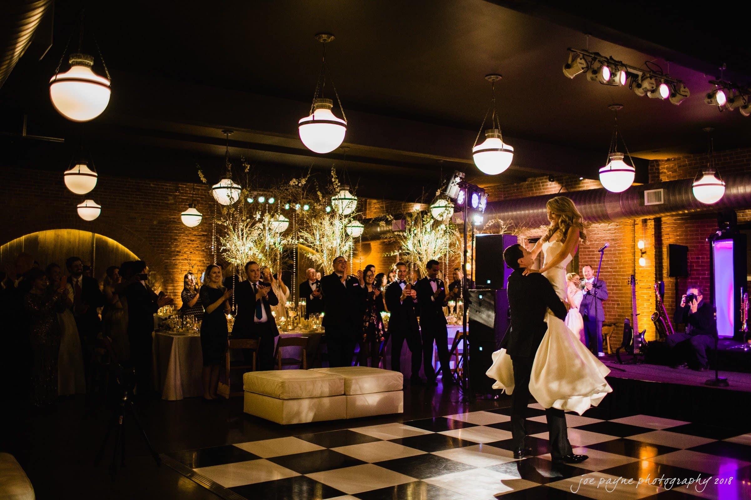 A joyful couple dances on a checkered floor at a warmly lit reception. The groom lifts the bride, who wears a flowing dress. Guests stand around clapping and smiling amidst elegant decor with soft lighting and hanging lights.