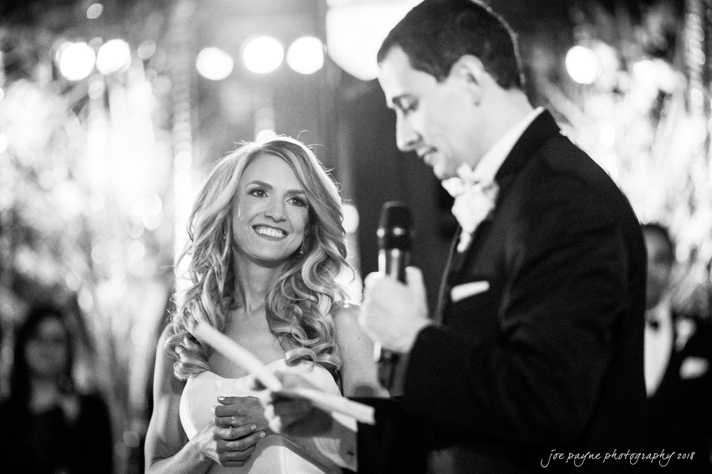 A bride and groom at their wedding. The groom is reading from a paper while holding a microphone, and the bride is smiling at him. The scene is black and white, with guests visible in the blurred background.
