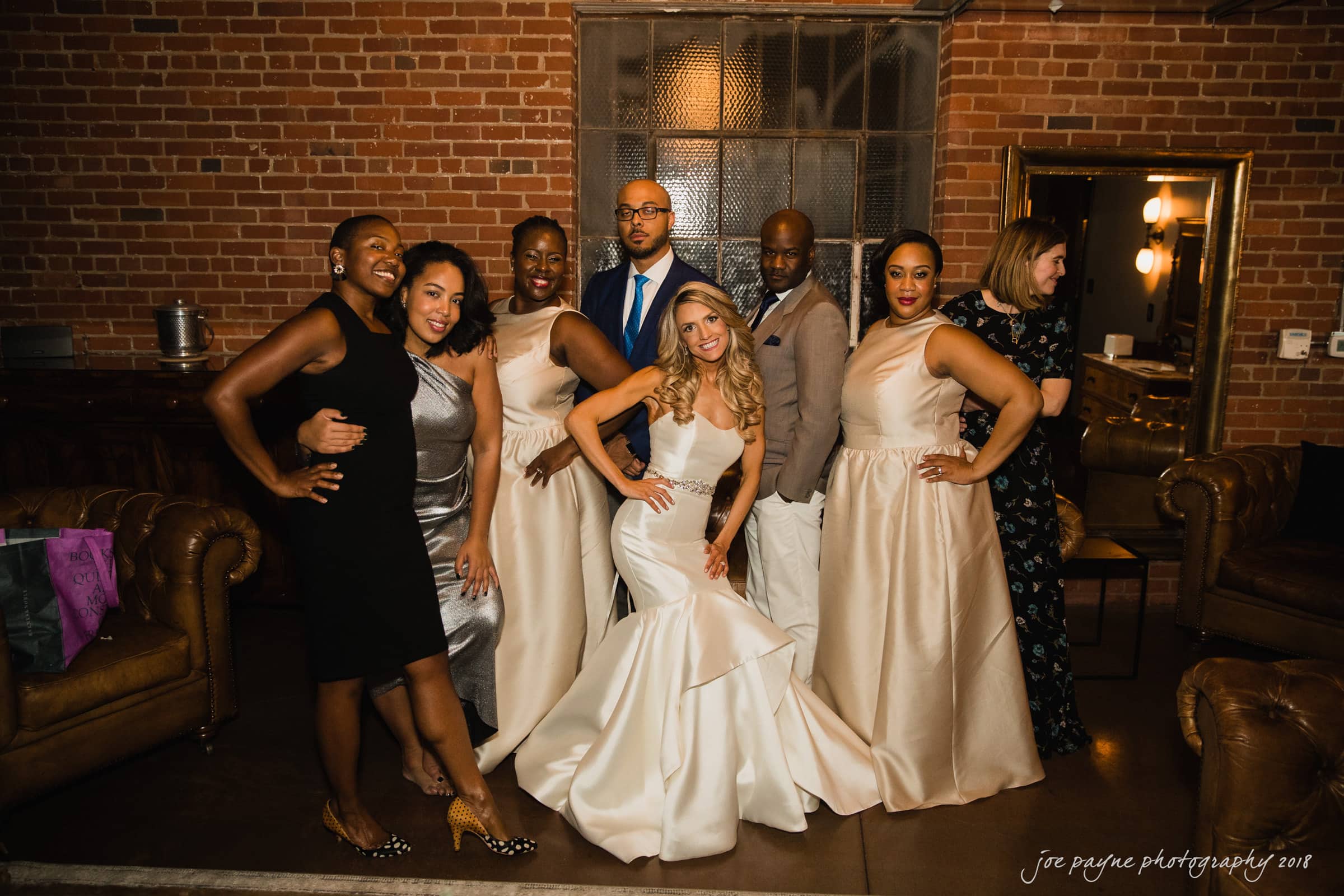 A group of people posing indoors in formal attire. Several women are wearing elegant dresses, and the men are in suits. They are standing in a room with exposed brick walls and leather chairs, smiling and striking playful poses.