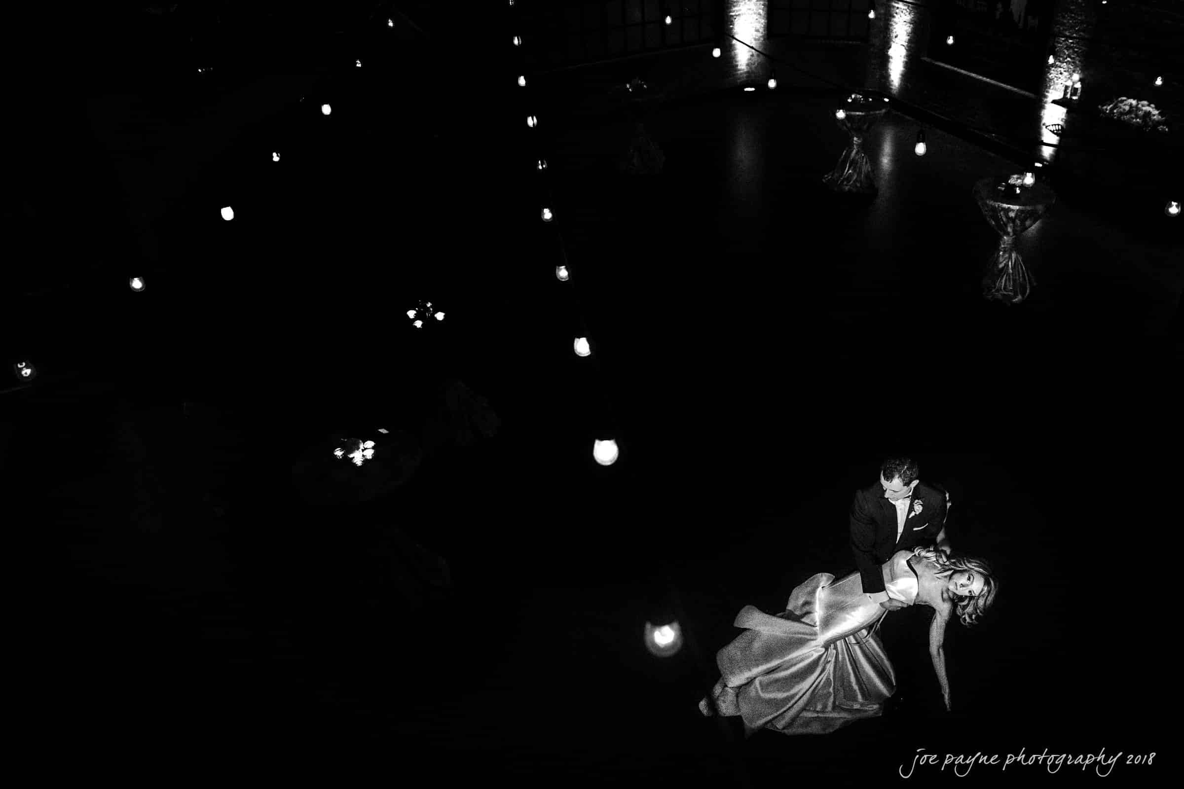 A couple in formal attire is dancing under dim lighting in a mostly dark room. She is being dipped gracefully by her partner. Candle-like lights line the floor, creating a romantic ambiance.