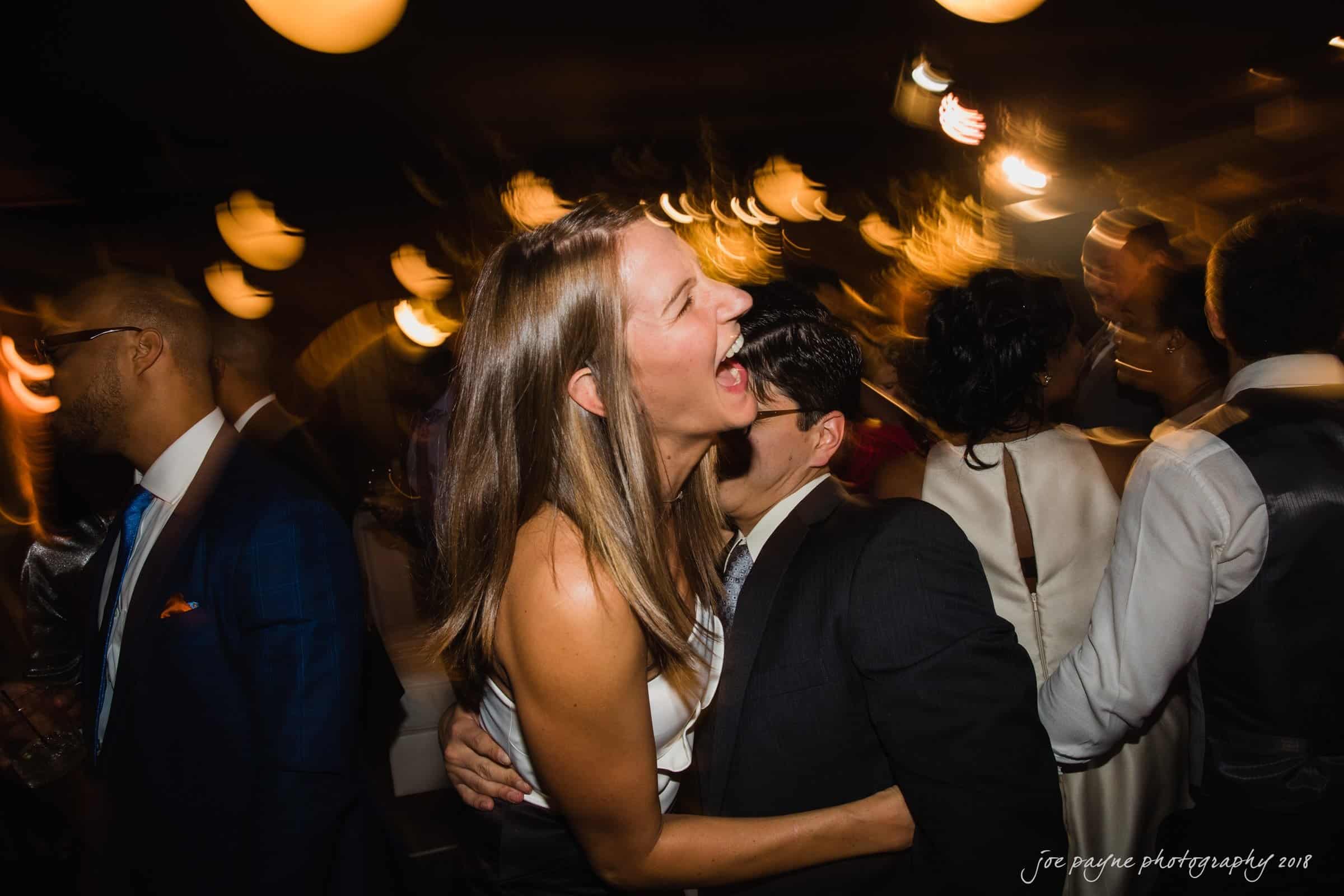 A joyful woman with long hair laughs while embracing a person in a suit on a dance floor. The background is blurred, capturing the dynamic atmosphere and movement of the celebration.