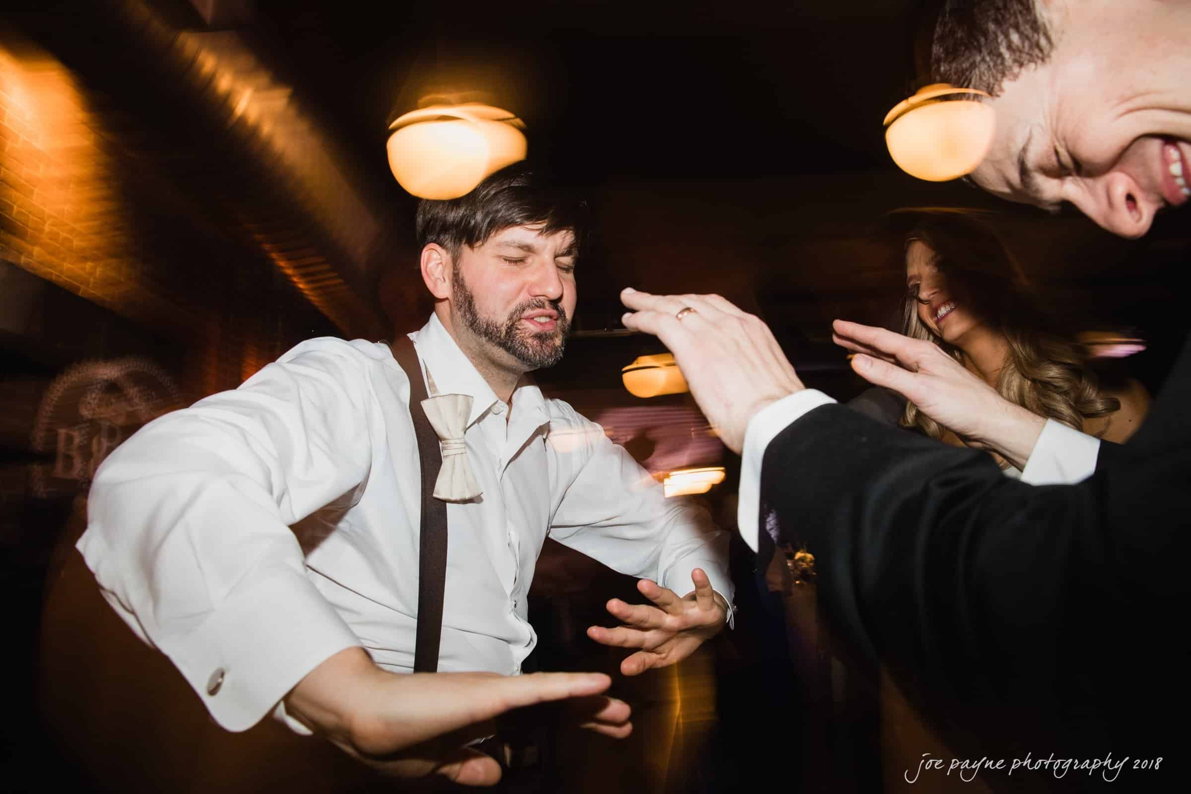 A man in a bow tie dances energetically under warm lighting. His expression is focused and joyful, with blurred motion conveying movement. A woman beside him also dances, smiling with hands raised. The atmosphere is lively and celebratory.