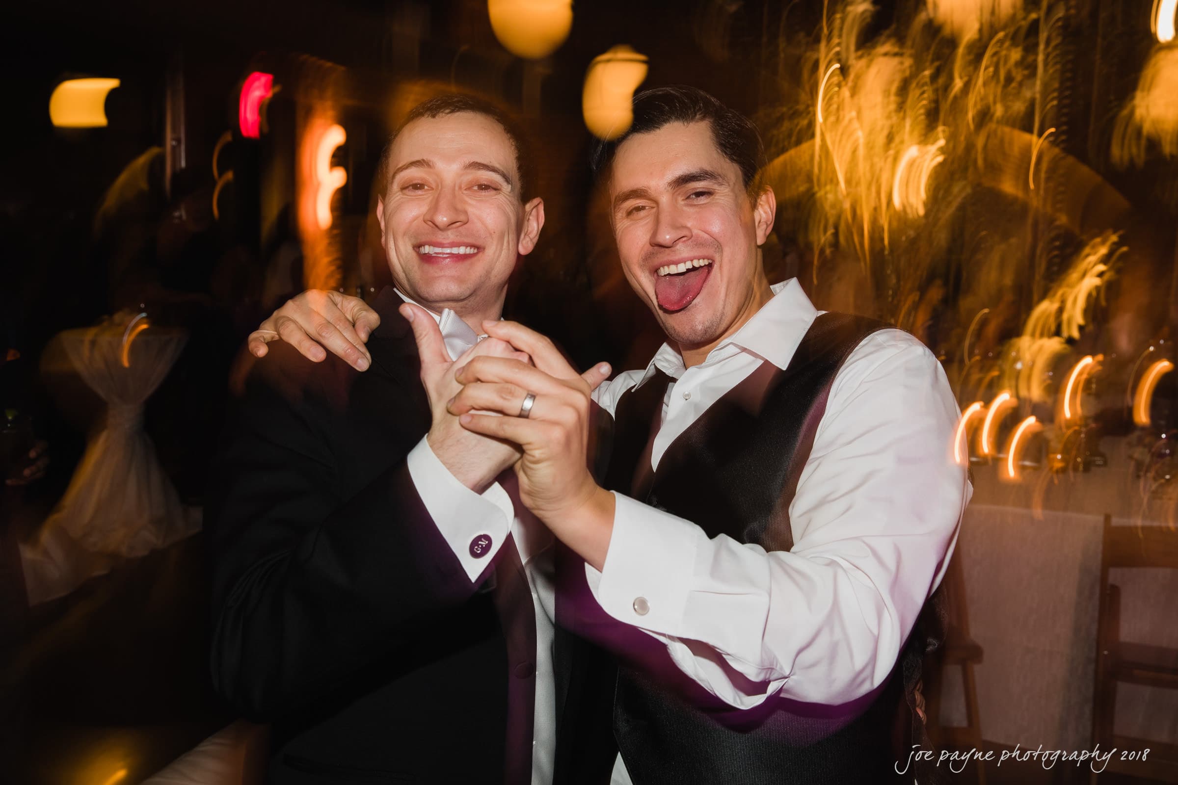Two men in formal wear are smiling and holding hands while dancing at an indoor event. The background features blurred lights, giving a lively and festive atmosphere.
