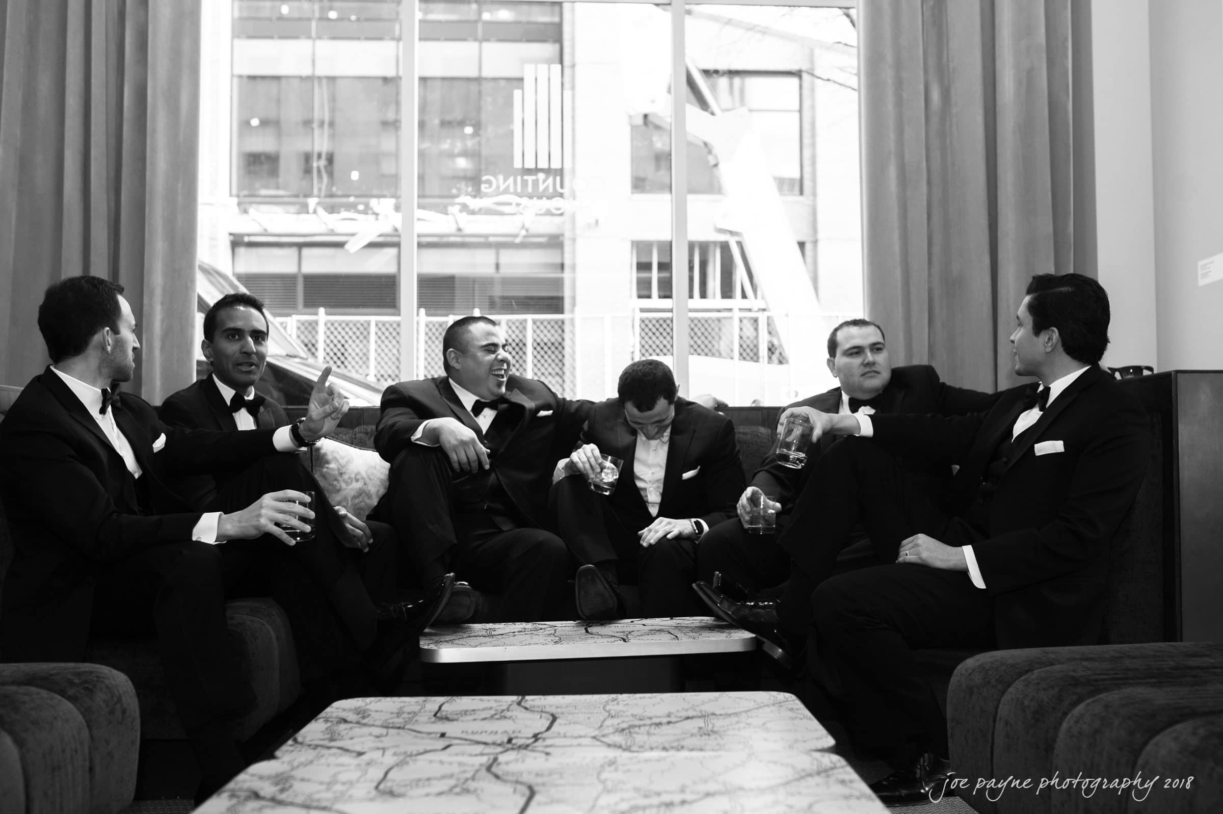 A group of six men in formal suits are sitting and chatting on sofas in a lounge area. They are holding drinks and appear engaged in conversation, with a large window and a view of a building in the background. The photo is in black and white.