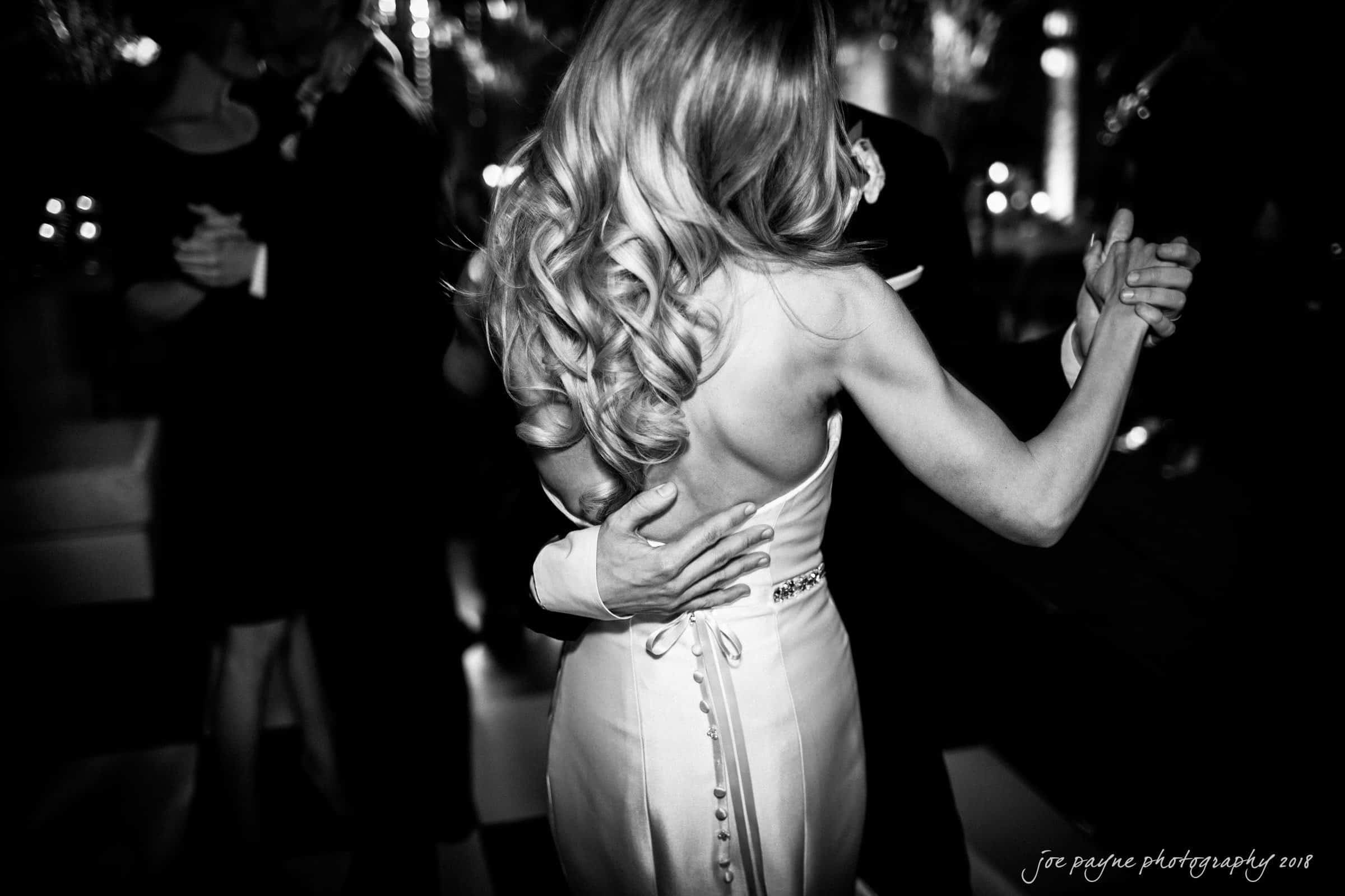 A couple elegantly dances at an event. The woman has long, wavy hair and wears a backless dress, while the man is in a suit. The black-and-white setting creates a timeless and classic atmosphere.