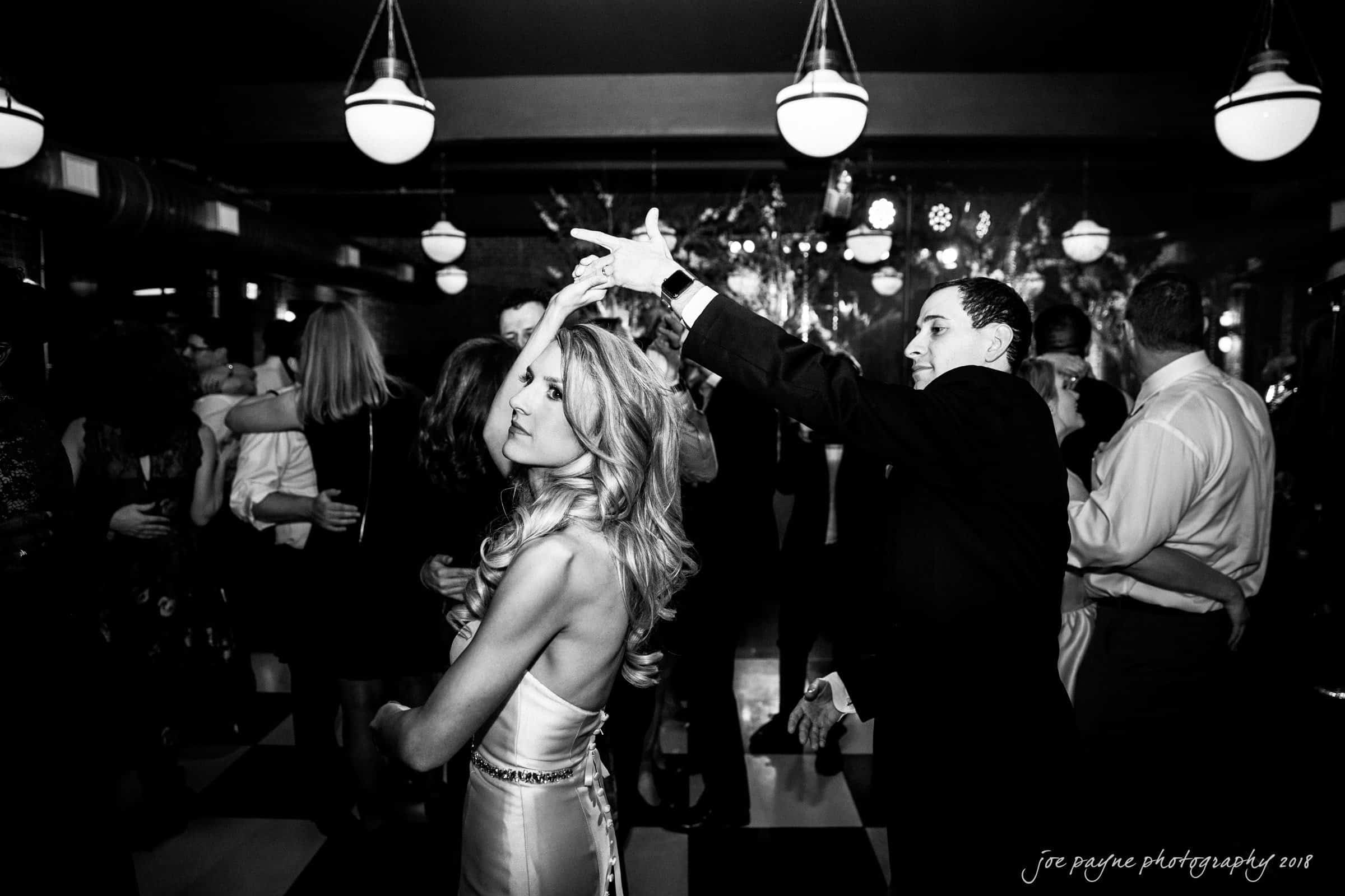 A couple dances elegantly on a checkered floor in a dimly lit room. The woman, wearing a sleeveless satin dress, twirls gracefully. People are gathered in the background, adding to the lively atmosphere.