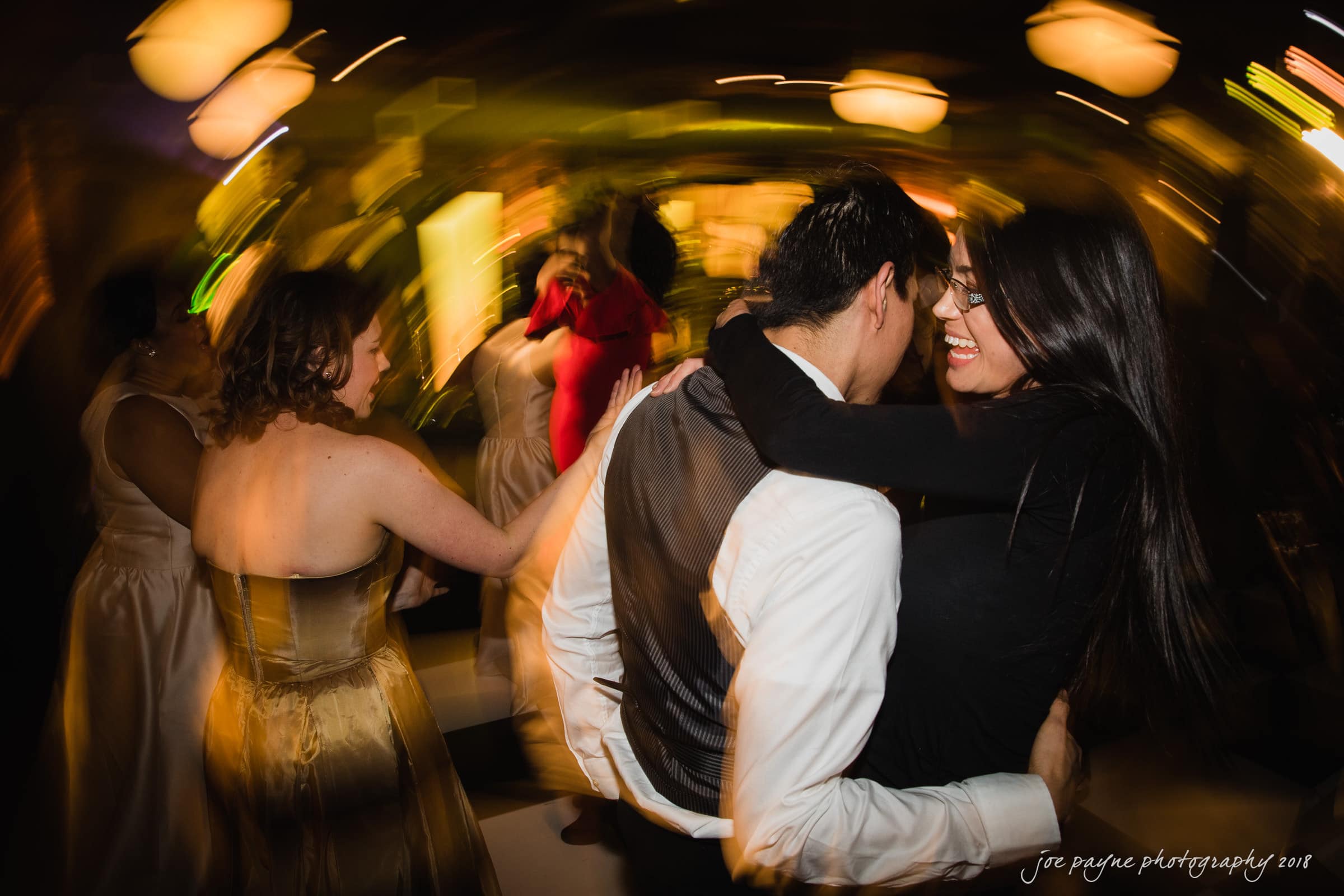 A couple dances closely, smiling at each other amid a lively blurred background. People in elegant attire move around them under warm lighting, creating a vibrant, festive atmosphere.