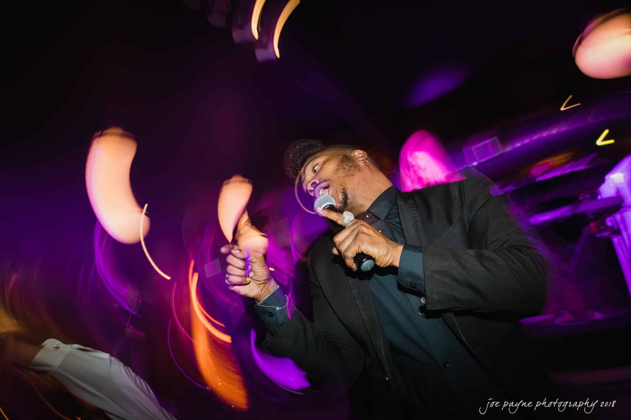 A man in a black suit sings passionately into a microphone on stage, surrounded by swirling purple and orange lights. The vibrant motion effect suggests an energetic performance.