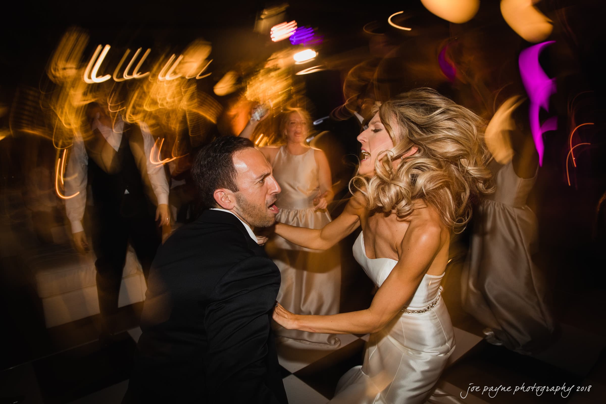 A couple energetically dancing at a wedding reception. The woman, in a white dress, has flowing hair, and the man is in a dark suit. The background shows other guests in motion, and the atmosphere is lively with colorful lights.