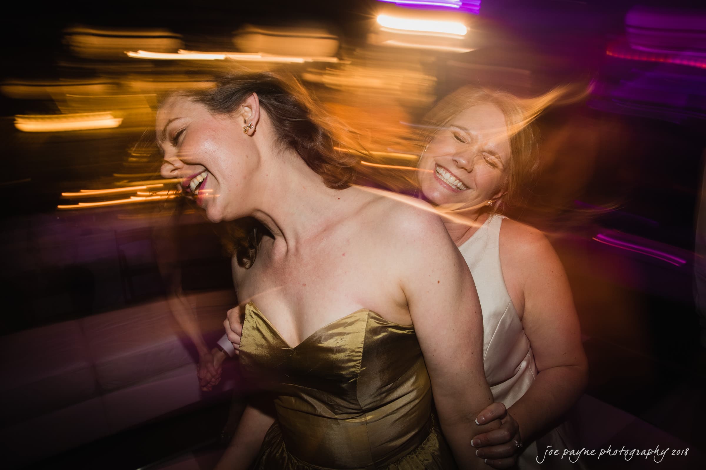 Two women dancing joyfully, with blurred motion capturing their movement. One wears a golden dress and the other a white outfit. Both have big smiles, and the background is filled with colorful lights.