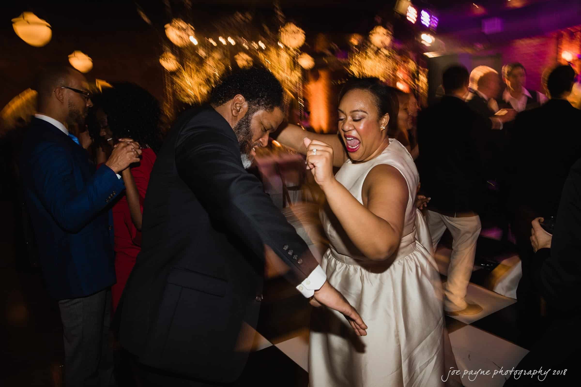 A joyous couple dances energetically on a dimly lit dance floor, surrounded by other partygoers. The woman wears a white dress and the man a black suit. The warm lighting and lively atmosphere suggest a celebratory event.
