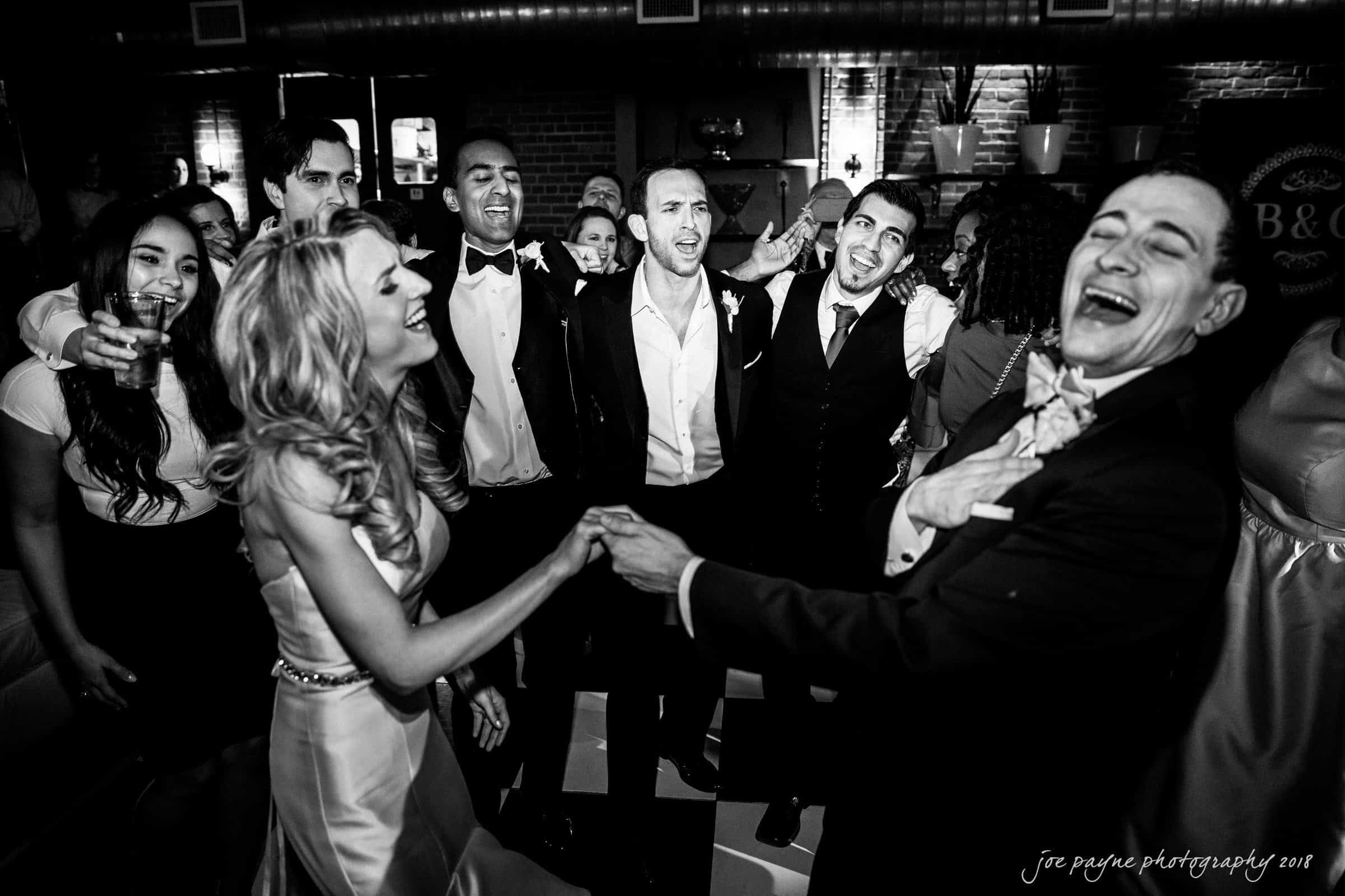 A joyful black-and-white scene of a wedding reception, with a group of people dressed in formal attire singing and laughing together on the dance floor. A bride in a gown leads one side of the circle, holding hands with a guest.