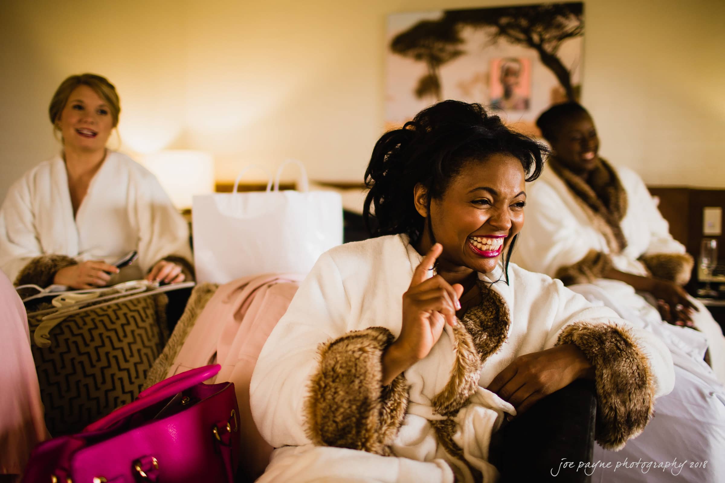 Three women in cozy robes sit in a warmly lit room, smiling and enjoying themselves. One woman in the foreground is laughing with her hand raised. A bright pink bag sits nearby on a bed. The atmosphere is joyful and relaxed.