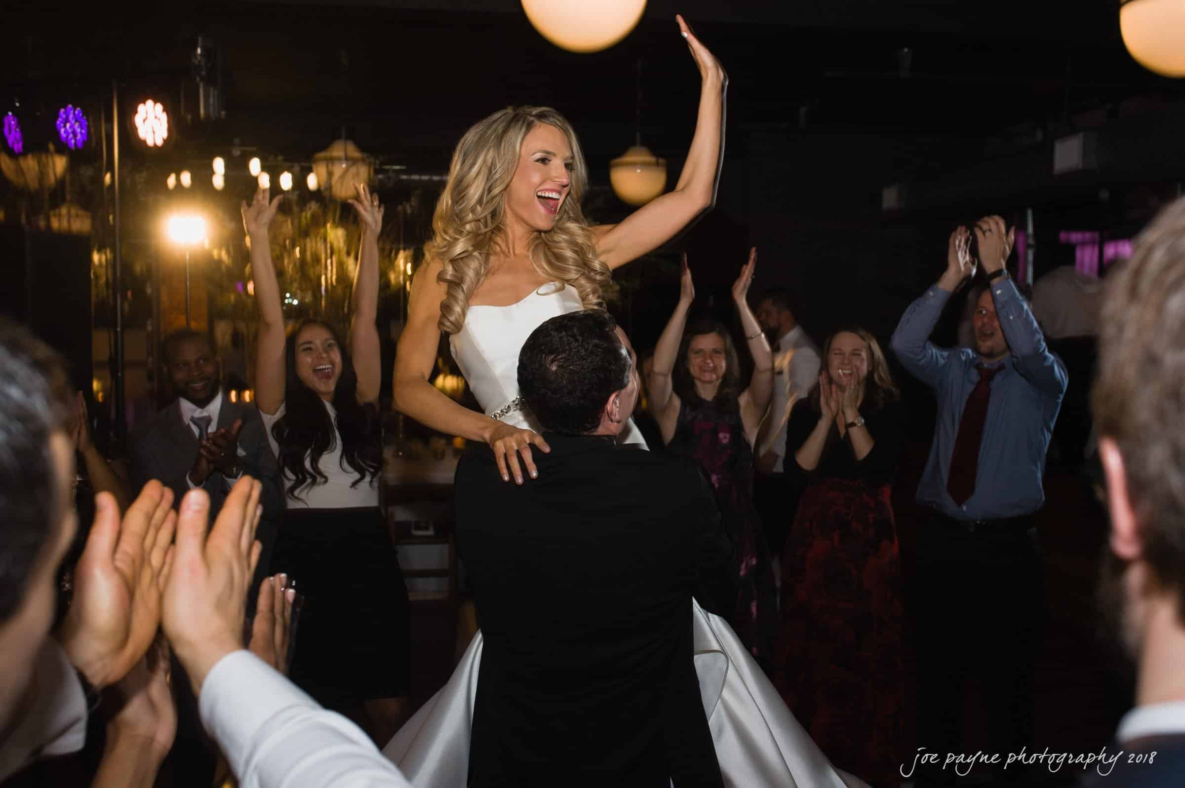 A bride in a white gown is joyfully lifted by a man in a suit at a wedding reception. Guests around them are clapping and smiling, capturing a moment of celebration and happiness.