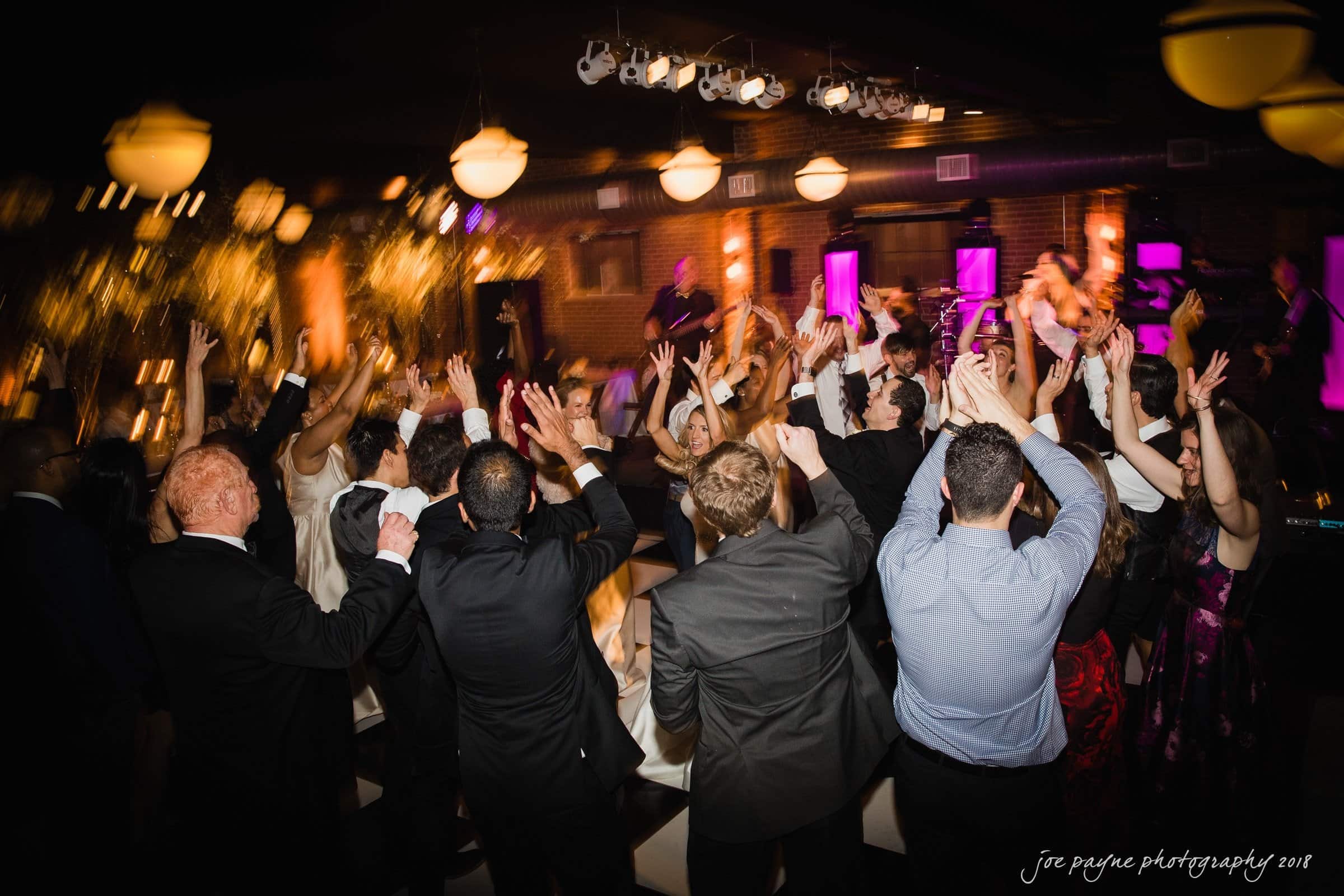 A lively group of people dancing at a party, with their hands in the air. The ambiance features dim lighting and colorful effects. The setting suggests a celebration or festive event, with attendees dressed in formal wear.