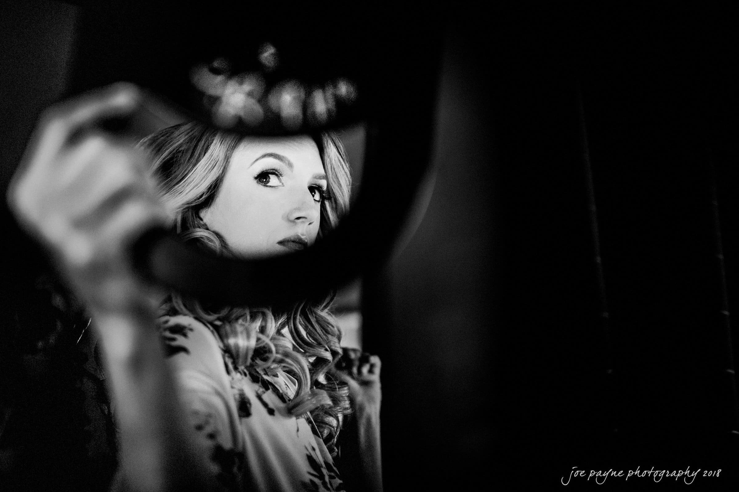 Black and white image of a person with long hair looking into a round mirror, holding it with their left hand. The focus is on their face and reflection, with soft lighting creating a dramatic and intimate atmosphere.