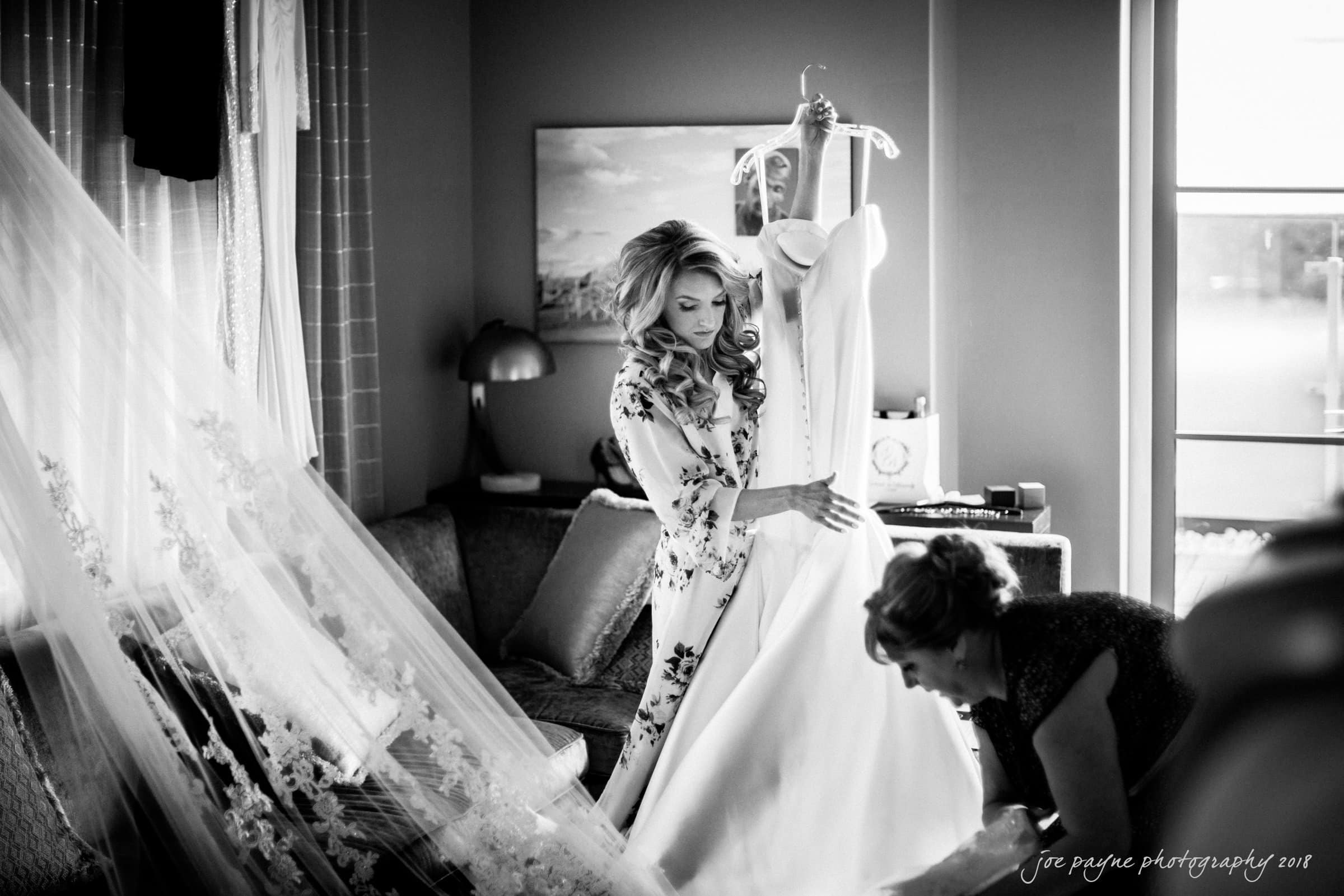 A bride-to-be in a floral robe holds her wedding dress next to a large window. A long veil with lace details hangs nearby. Another woman, possibly assisting, leans forward arranging the dress. The room has a cozy, elegant ambiance.