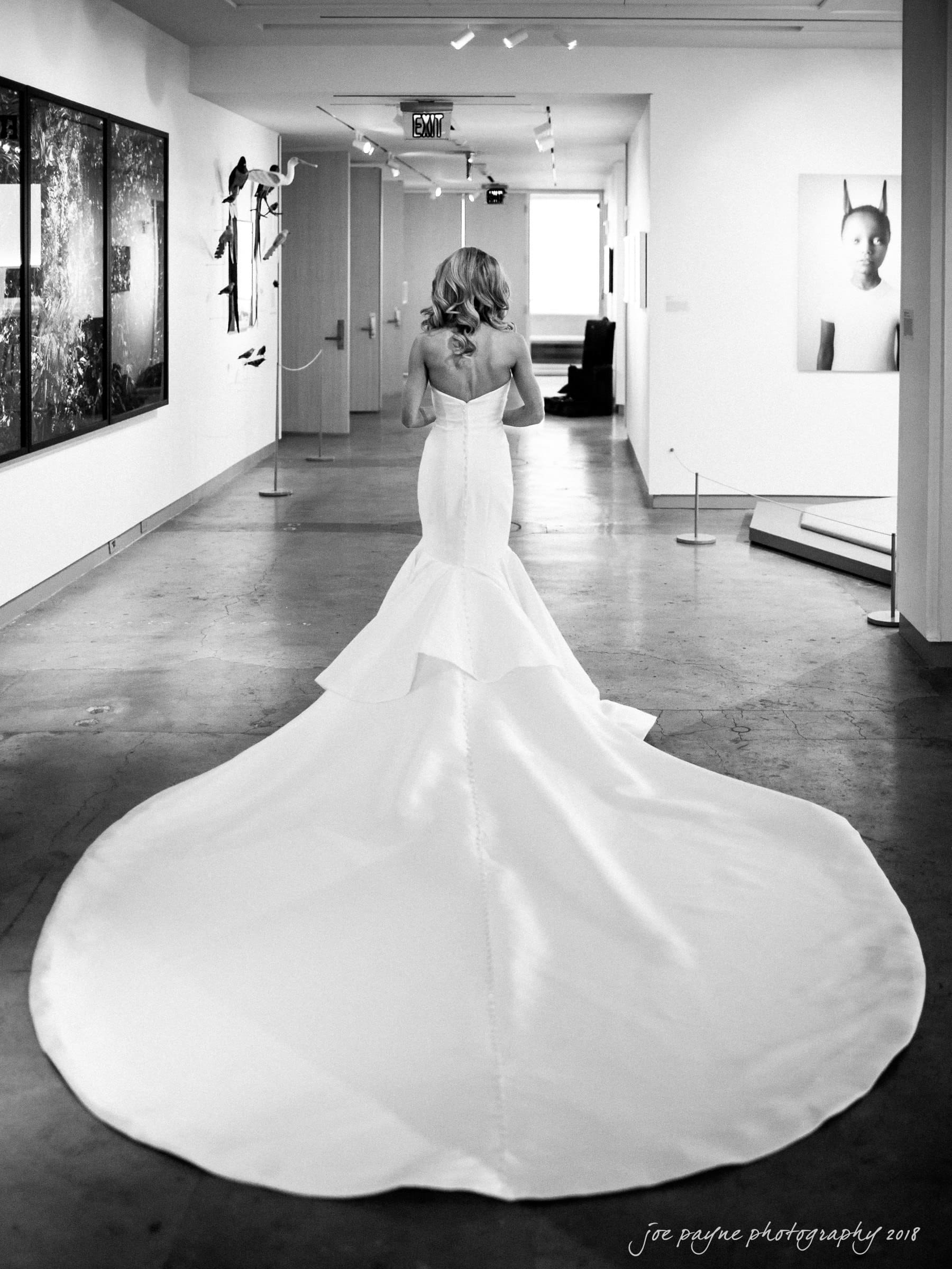 A woman in a long, elegant wedding dress with a flowing train walks down a corridor adorned with art. The setting is minimalistic and modern, with light streaming in from a window at the end.
