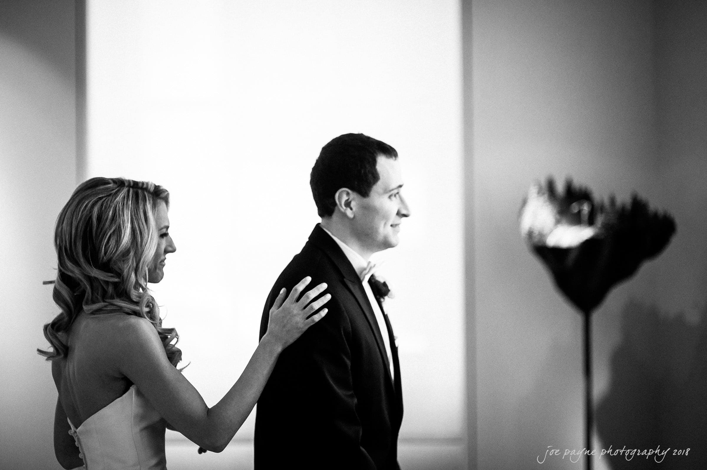 A bride and groom smile warmly as they stand close together. The bride rests her hand gently on the grooms shoulder. The background is softly lit, creating an intimate, joyful atmosphere.