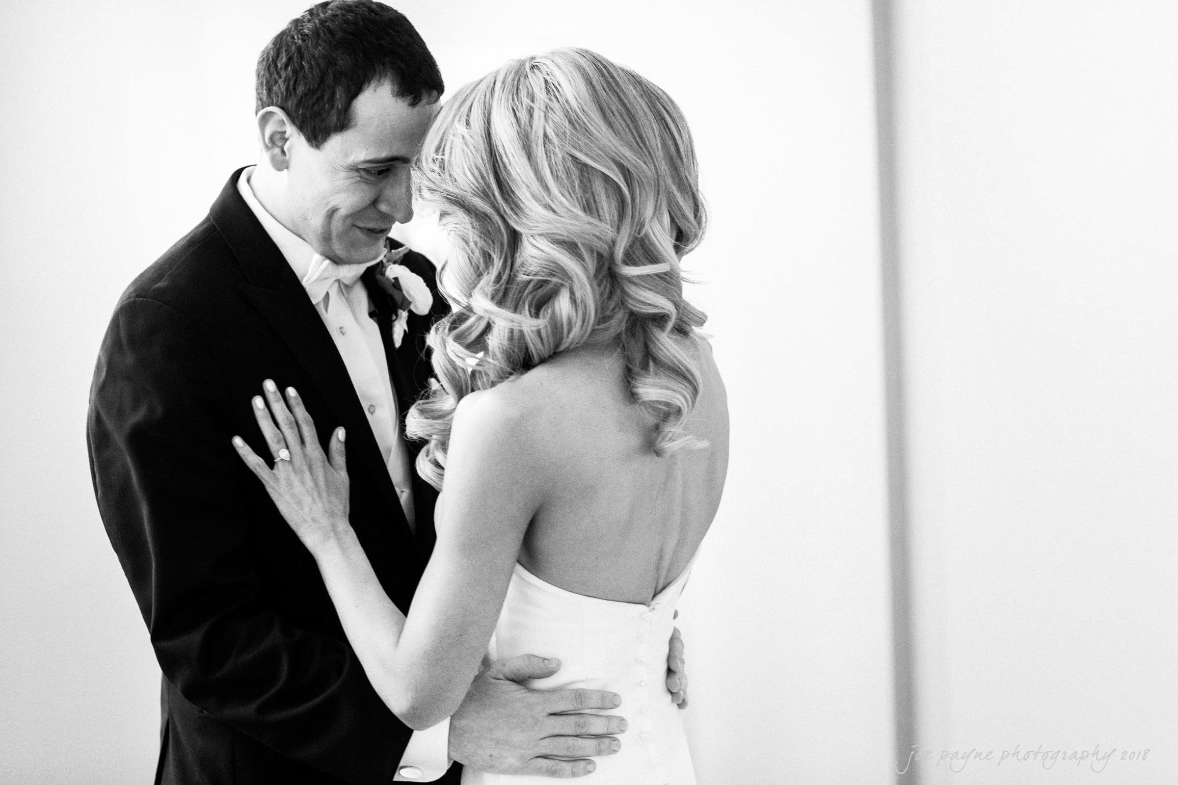 A black and white photo of a bride and groom embracing. The groom is in a suit with a boutonniere, and the bride is in a strapless dress with long, wavy hair. They are smiling and touching foreheads, creating an intimate and joyful moment.