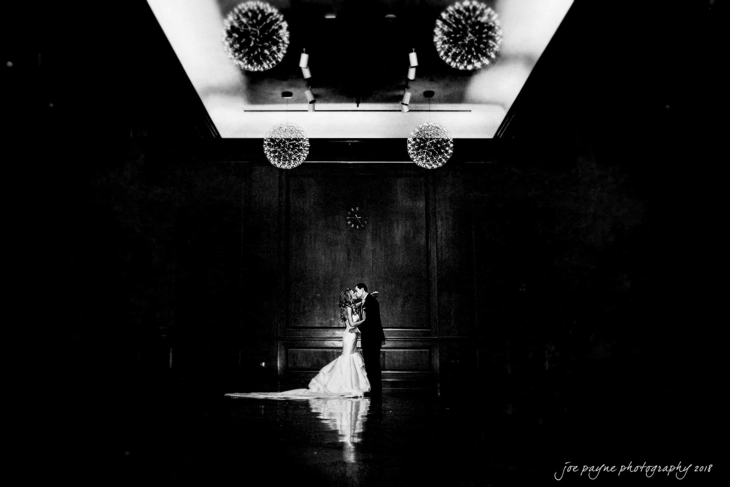 A bride and groom share an intimate moment in the center of a dimly lit room with elegant hanging chandeliers, reminiscent of a Duke Chapel wedding. The atmosphere is romantic and dramatic, emphasizing their connection.