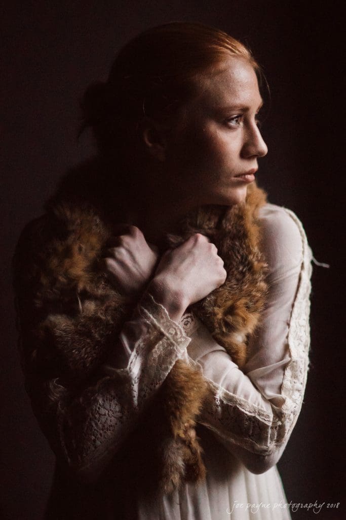 parker j pfister's french revolution workshop review A person with reddish hair stands in profile against a dark background, wearing a lace dress and holding a fur stole around their shoulders. The lighting casts soft shadows, emphasizing their contemplative expression.