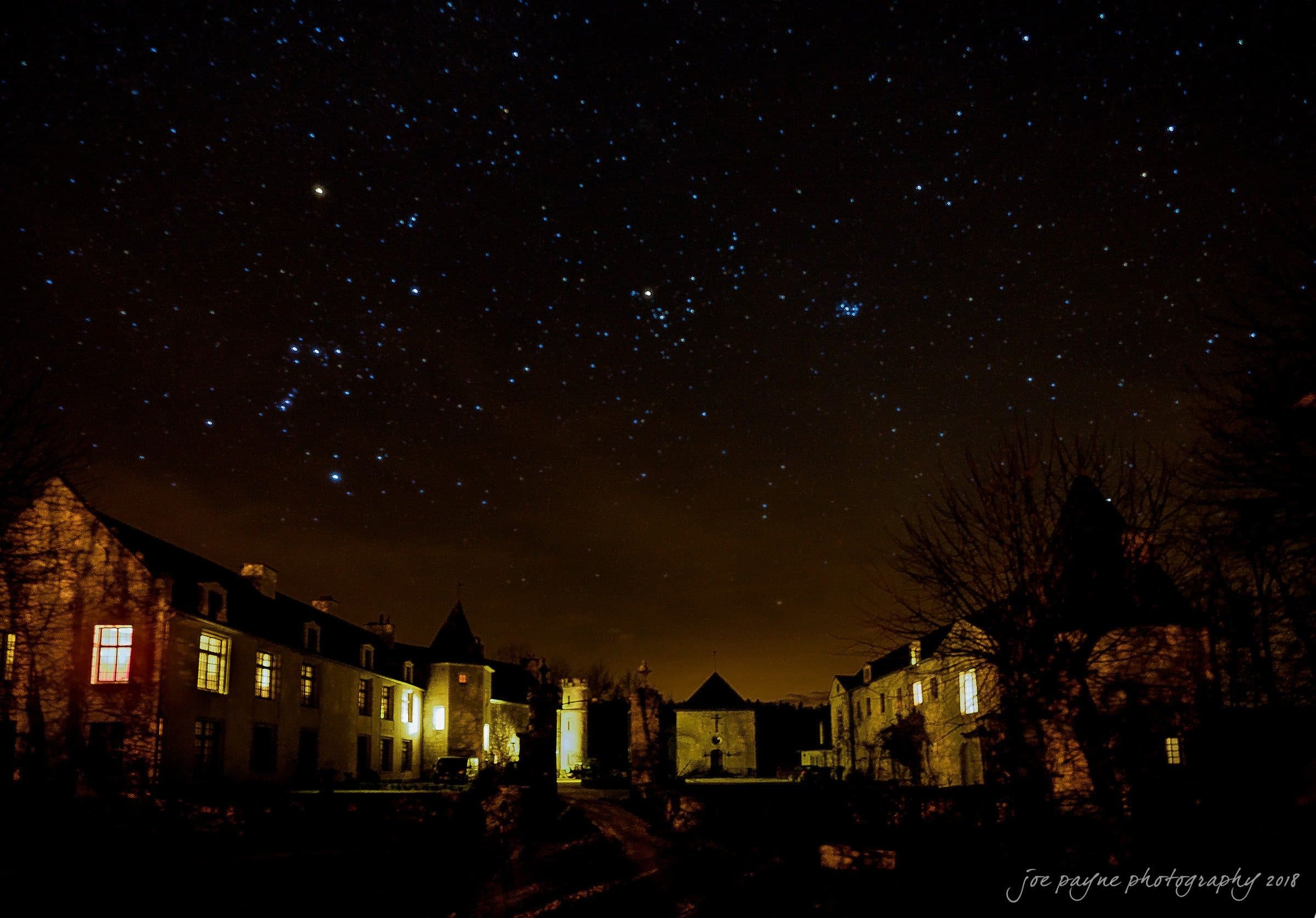 parker j pfister's french revolution workshop review A starry night sky over a row of historic stone buildings. Warm lights illuminate the windows, creating a cozy contrast to the dark, expansive sky filled with constellations.