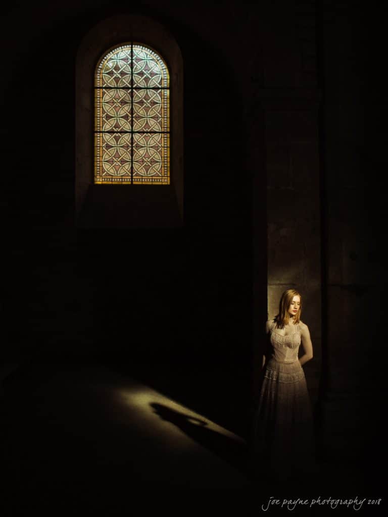 parker j pfister's french revolution workshop review A woman in a white dress stands in a dimly lit room, illuminated by soft light from a stained-glass window. Shadows fall around her, creating a serene and mysterious atmosphere.