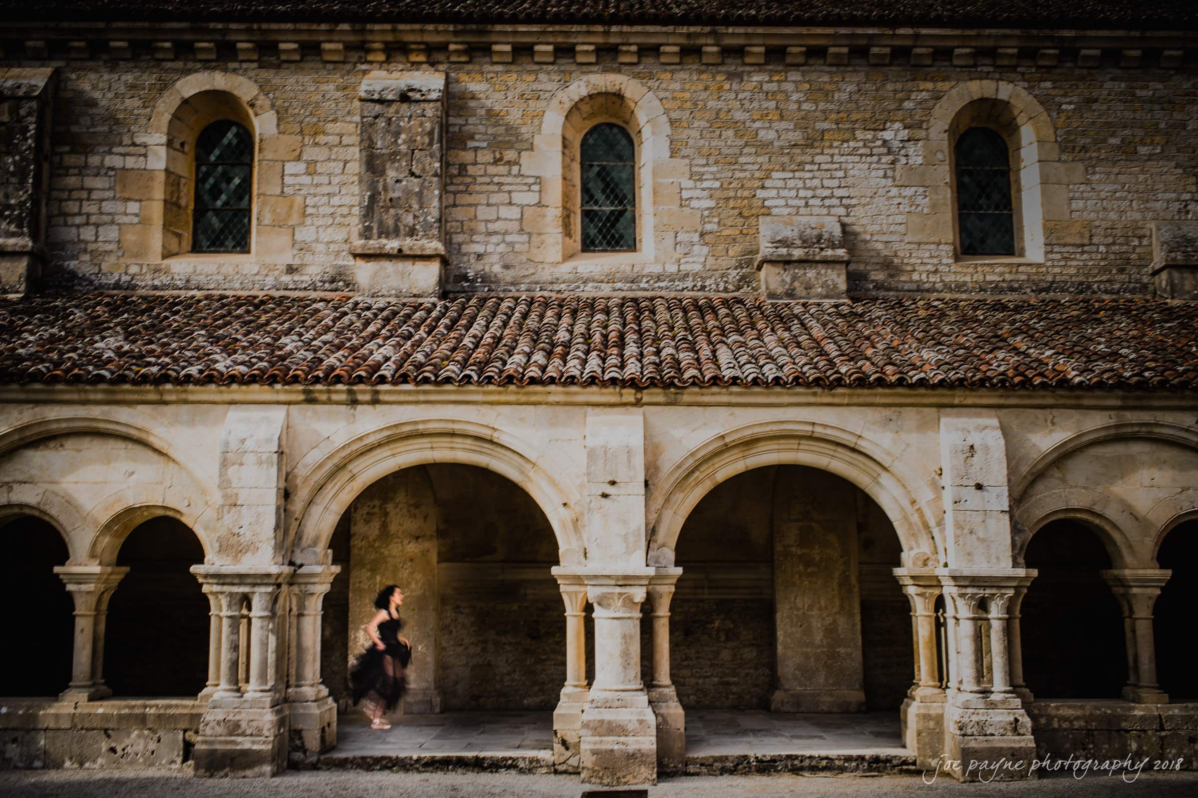 parker j pfister's french revolution workshop review A historic stone building with arched columns and a tiled roof. A person in a black dress walks through the arches. The structure has small windows with a rustic appearance, creating a serene, ancient atmosphere.
