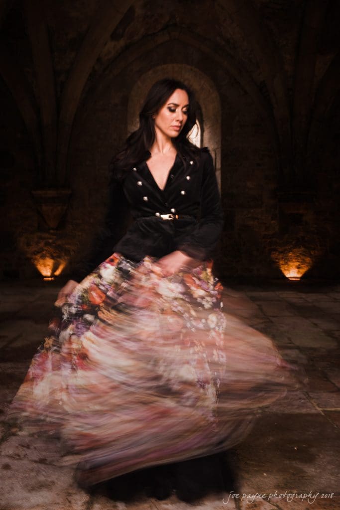parker j pfister's french revolution workshop review A woman in a dark jacket twirls in a dimly lit stone hall, creating a blurred effect with her floral skirt. The arching stone ceiling adds an ancient ambiance, and warm lights illuminate the walls.