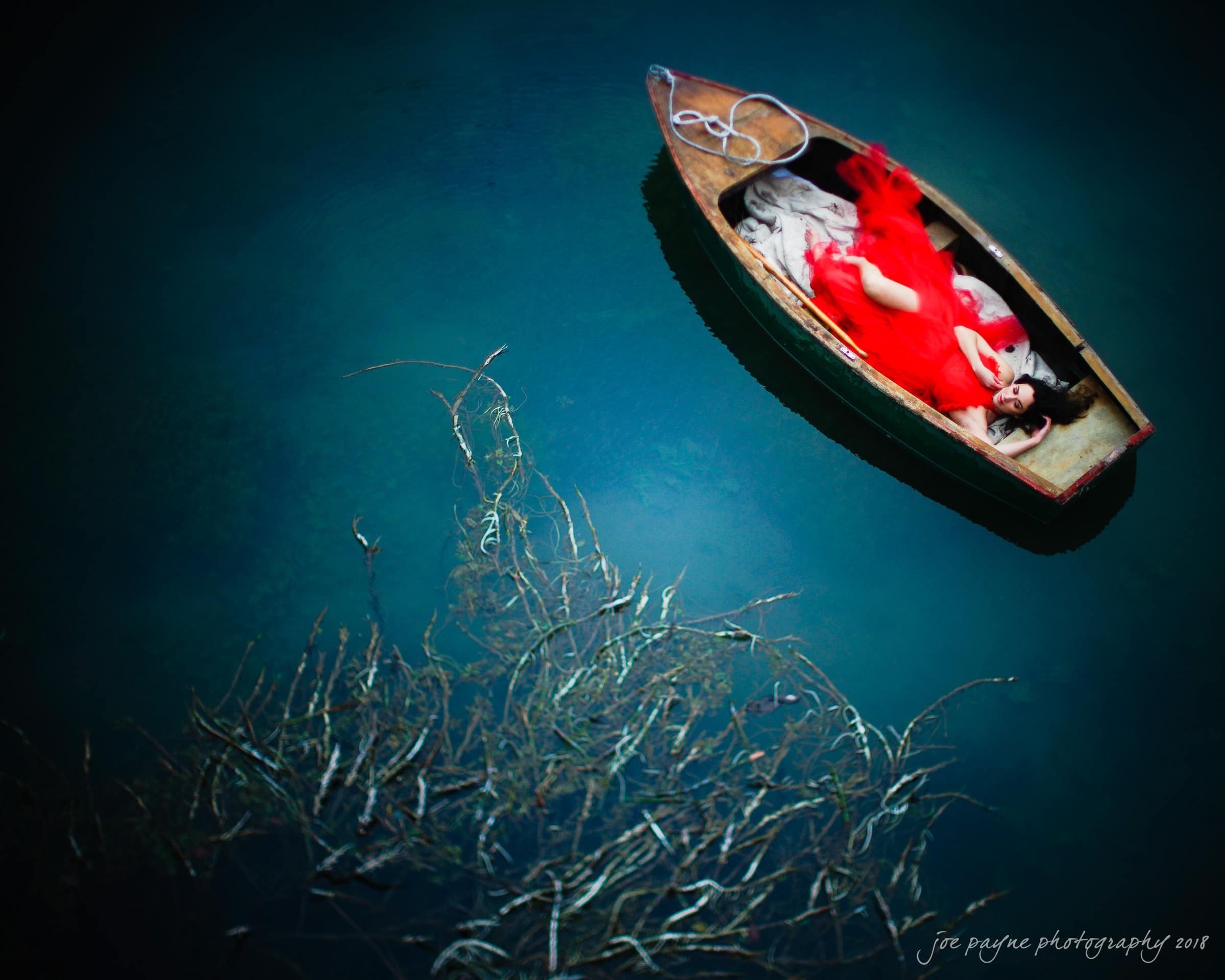parker j pfister's french revolution workshop review A woman in a flowing red dress lies in a small wooden boat floating on calm, dark blue water. She is surrounded by submerged, leafless branches. The scene conveys a serene and dreamy atmosphere.
